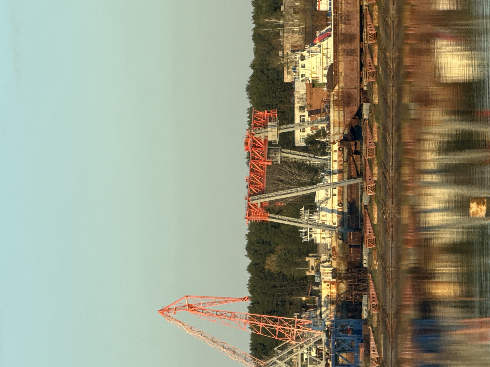 The image depicts the Kama shipyard in Chaikovsky, Russia, featuring orange industrial cranes along the waterfront. The foreground shows a clear sky, contrasting with the detailed elements of the shipyard. The scene includes waterfront buildings, machinery, and reflections on the water, creating a sense of depth and industrial activity.