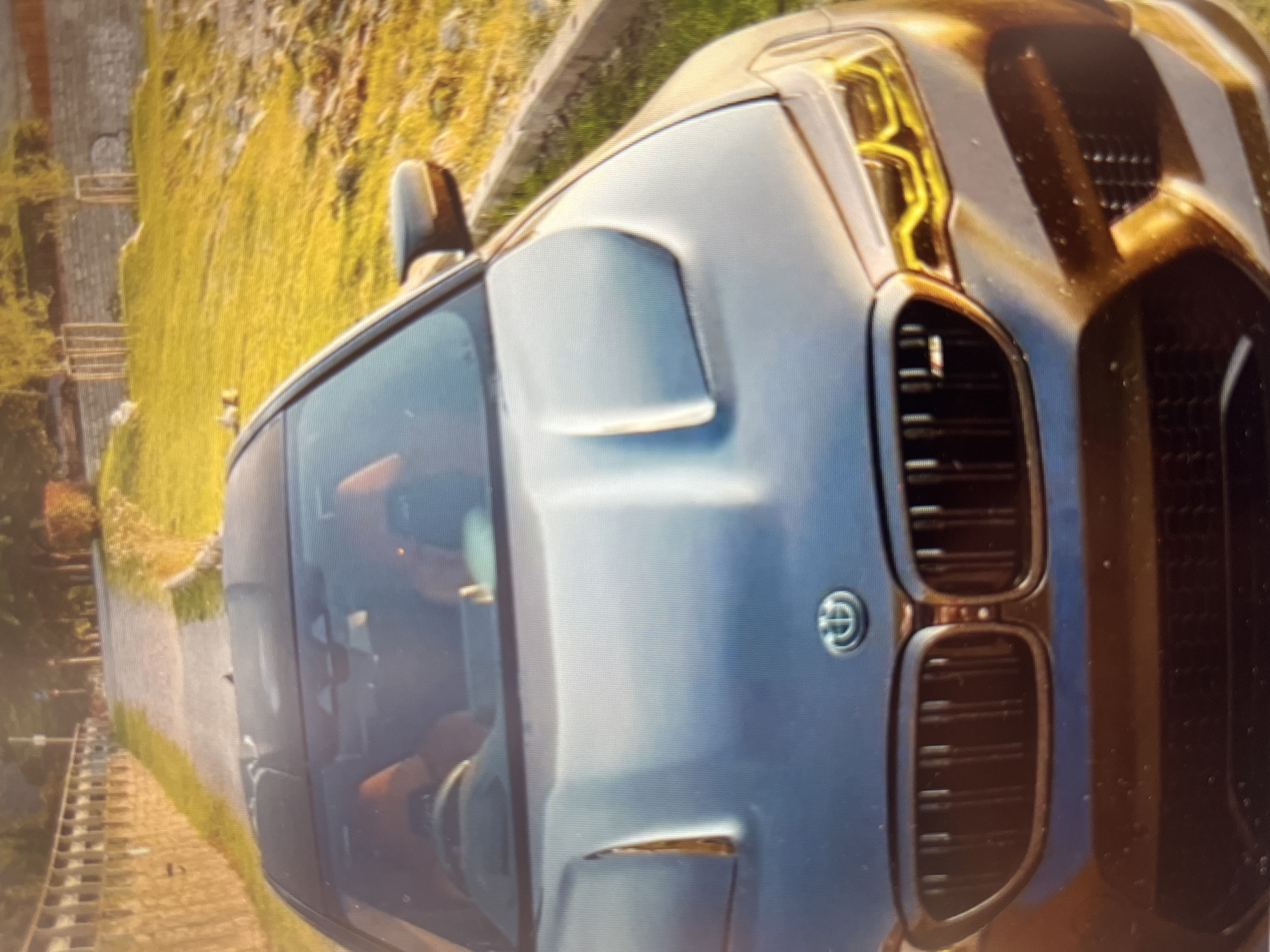 An upward-angled, slightly blurred, and rotated photograph captures the front-right side of a modern dark-colored SUV or large car, likely a dark blue or grey, under bright daylight. The vehicle features prominent yellow LED daytime running lights that create a distinctive "eyebrow" effect above the headlights. Its large, vertical kidney grilles, a characteristic design element, are visible on the front, with a circular emblem situated between them, though the specific details of the emblem are indistinct due to blur.
A driver is partially visible through the tinted front-right passenger window and windshield. The car appears to be on a narrow road or path, possibly in a rural or mountainous setting. In the background, lush green foliage covers a hillside or embankment, and a stone wall or old structure is visible on the left side of the frame. The overall impression is of a car driving or parked in a scenic, natural environment. The image quality suggests it might be a photo taken of a screen.