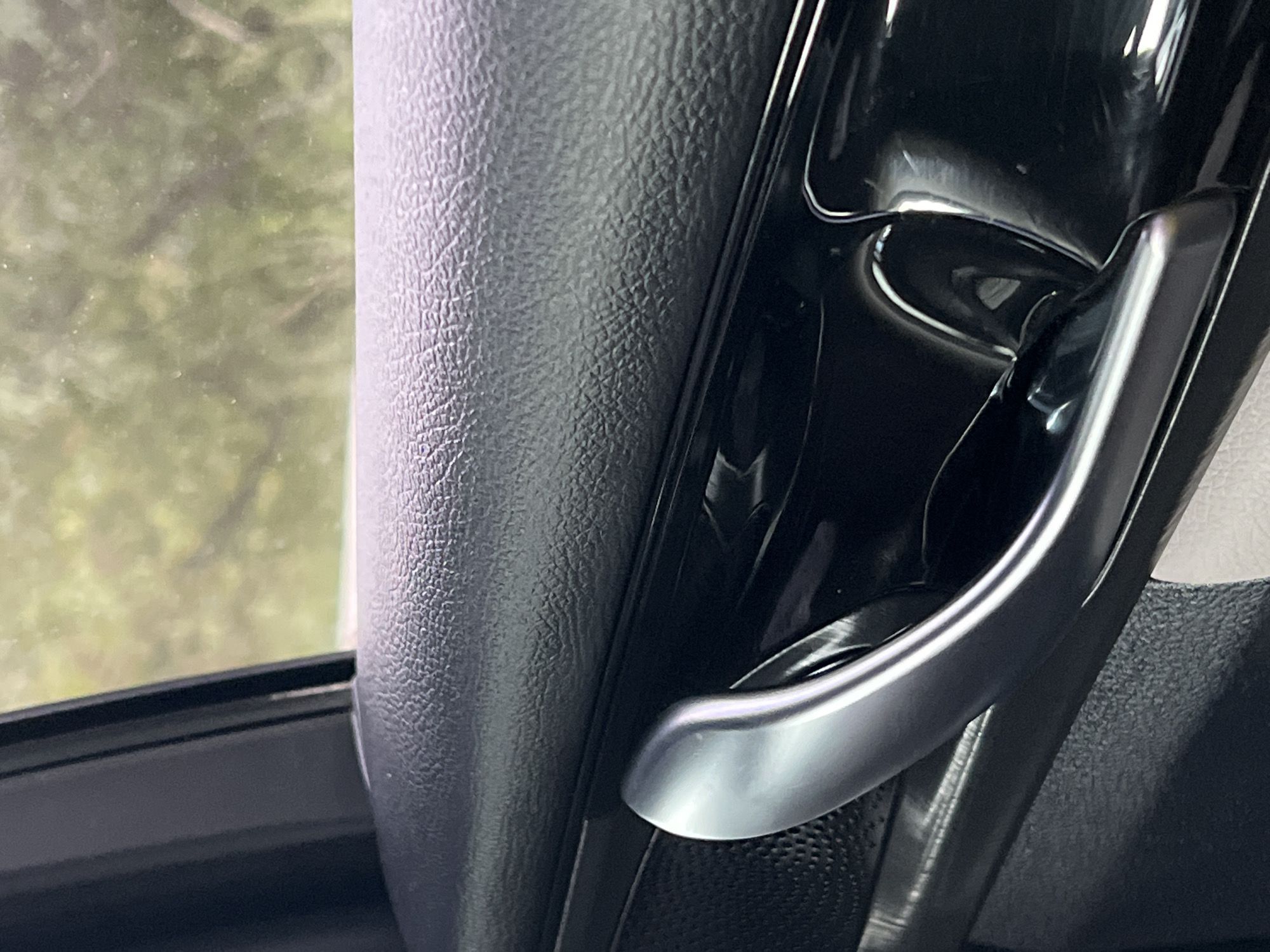 The image shows a close-up of the interior of a vehicle, focusing on a door handle. The door handle is made of a silver-toned metal and has a curved, ergonomic shape. It is mounted on a black, textured surface that appears to be part of the car's door panel. Above the handle, a portion of a window is visible, through which out-of-focus green foliage can be seen, suggesting the vehicle is outdoors. The lighting is bright, highlighting the reflective surfaces of the handle and the textured pattern of the door panel. There is no indication of the vehicle's location in Yekaterinburg, Russia, or any other specific geographical information. The scene is static and shows no people or actions.