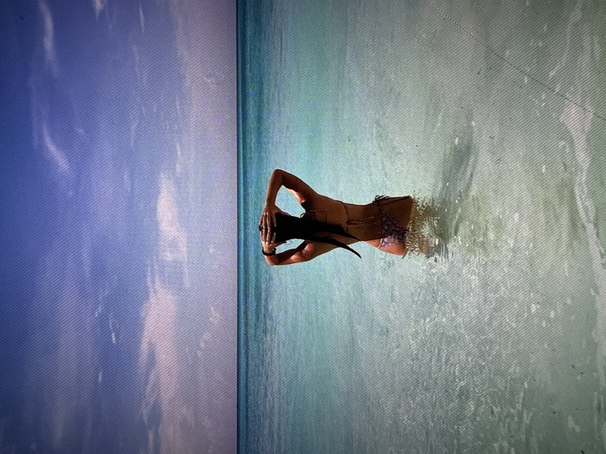 A woman in a bikini stands in the water, facing away from the camera, with her hands behind her head. She is positioned roughly in the center of the frame. The water appears shallow. The horizon line is visible in the background, separating the water from the sky. The sky is a light blue with some clouds. The location is a beach. The city and location are unknown.
