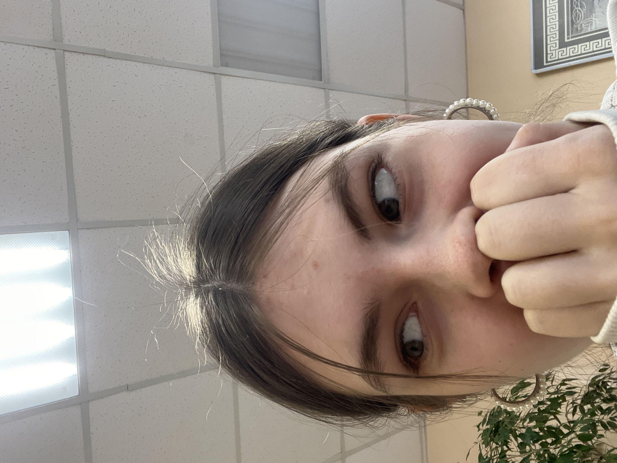 A low-angle photograph shows a young person with dark hair and brown eyes. The person's face is angled towards the camera, with their fingers partially covering their nose and mouth. They are wearing hoop earrings with small pearls. The background consists of a white, tiled ceiling with fluorescent lights and part of a framed picture with a classical Greek key design. A green leafy plant is also visible in the lower right corner. The location of Saint Petersburg, Russia, cannot be confirmed from the image alone.