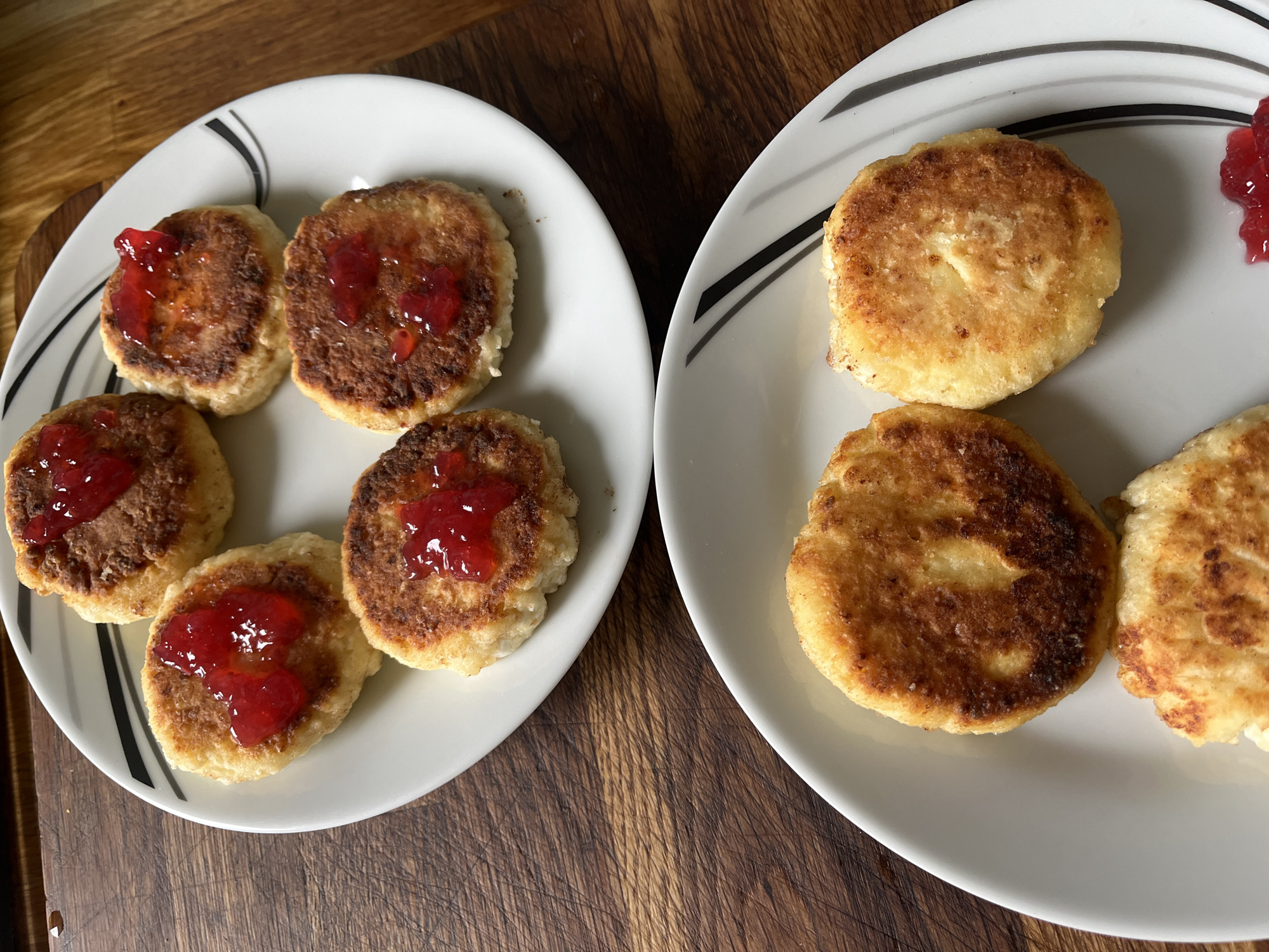 Two white plates filled with syrniki, a type of fried cheese pancake, are presented on a wooden cutting board. The top plate contains four syrniki, each topped with a dollop of red jam. Three of these syrniki are fully visible, while the fourth is partially obscured. The bottom plate holds three syrniki, with one almost completely visible and two partially visible. This plate lacks jam. The syrniki are golden brown and appear to be homemade. The lighting suggests an indoor setting. The exact location in the city of Unknown, Unknown cannot be determined from the provided image.