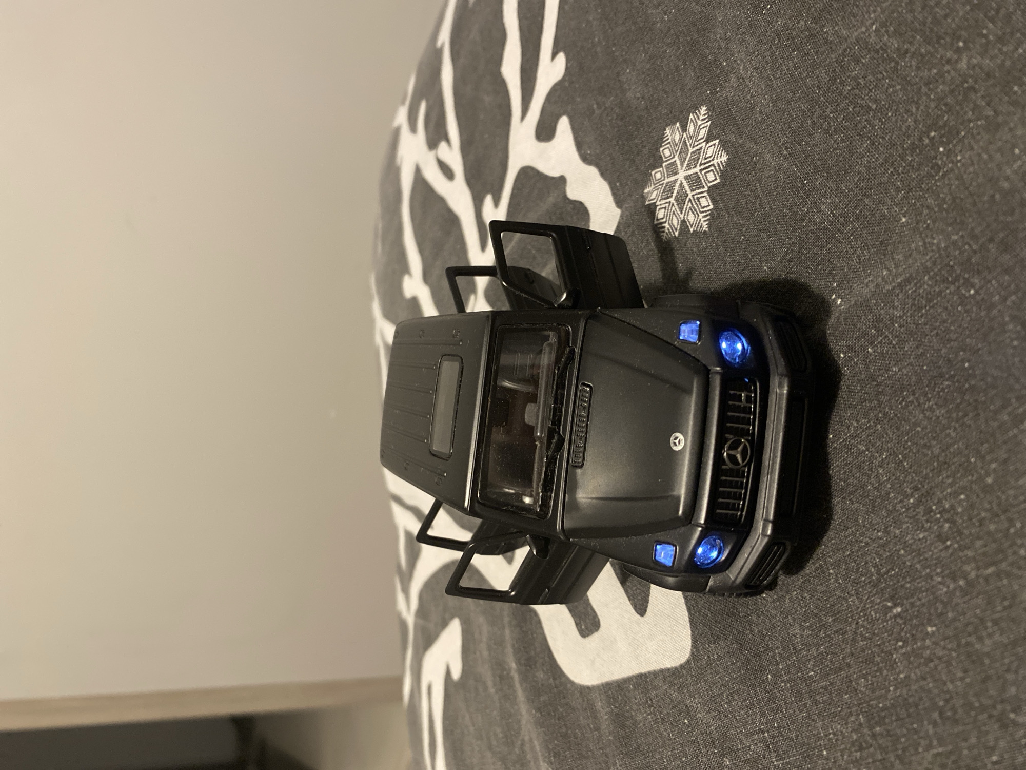 A black Mercedes-Benz G-Wagon toy car with its doors open is placed on a dark gray patterned surface. The car's headlights are illuminated with a bright blue light. The background consists of a plain, light-colored wall and a small portion of a doorway on the left side. The surface the car is on appears to be a fabric with a white, stylized tree-like or branch pattern, and a snowflake design in the lower right quadrant. The scene is indoors, and no individuals are present. The image does not contain any information that would allow for its location to be determined as Łódź, Poland.