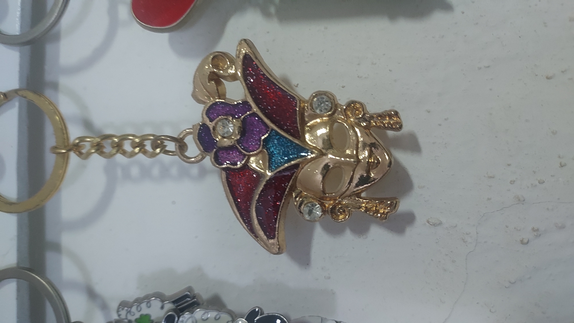 A close-up shot captures a gold-colored Venetian mask-shaped keychain with ornate details, hanging against a textured white wall. The mask itself is gold-toned and features a headdress adorned with red and purple enamel, accented by blue glitter. Two small, clear rhinestones are embedded on either side of the mask's eyes, and the mask has dangling gold charms. The keychain is suspended from a gold chain, which is attached to a gold keyring at the top of the frame. The wall exhibits a rough, uneven surface with some minor imperfections and a faint crack running vertically in the lower portion of the image. To the left, a portion of another keychain with a white and black sheep charm is visible, and to the right, a hint of a red heart-shaped object is present, suggesting other items are also displayed in the vicinity, possibly in a market or shop setting within Kogon City, Uzbekistan.