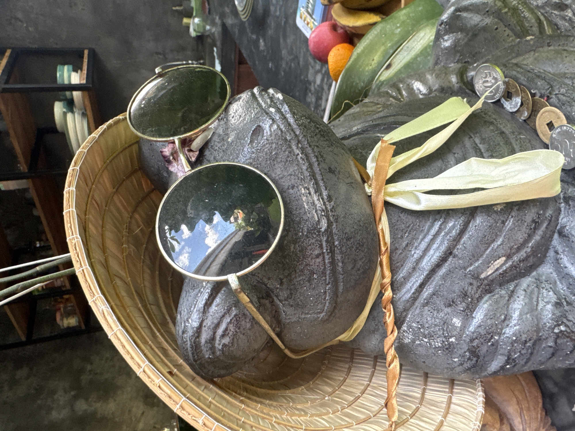 The image presents a close-up view of a dark, textured statue or carving, likely made of stone or a similar material. The statue's head is adorned with a pair of round, dark-lensed sunglasses. A traditional conical straw hat, characteristic of Southeast Asian regions, is positioned around the statue's head and neck.
Various ribbons and cords are draped over the statue's form, including light yellow, light green, and an orange-brown twisted ribbon. Several coins are strung together and rest on the statue's surface. Visible among these are modern Indonesian Rupiah coins, with "500 RUPIAH" and "BANK INDONESIA" clearly legible on one coin. Alongside them are older, corroded coins featuring square holes, indicative of ancient Chinese cash coins.
In the background, out of focus, details of a setting emerge. A wooden shelf holding stacked items, possibly plates or bowls, is partially visible. Further back, colorful fruits like a red apple and an orange can be seen nestled among green leaves. The bright lighting suggests a daytime setting. This arrangement, with its blend of traditional and playful elements, along with the specific coins and hat style, is consistent with the cultural context of Sukawati, Indonesia, known for its art and traditional crafts.