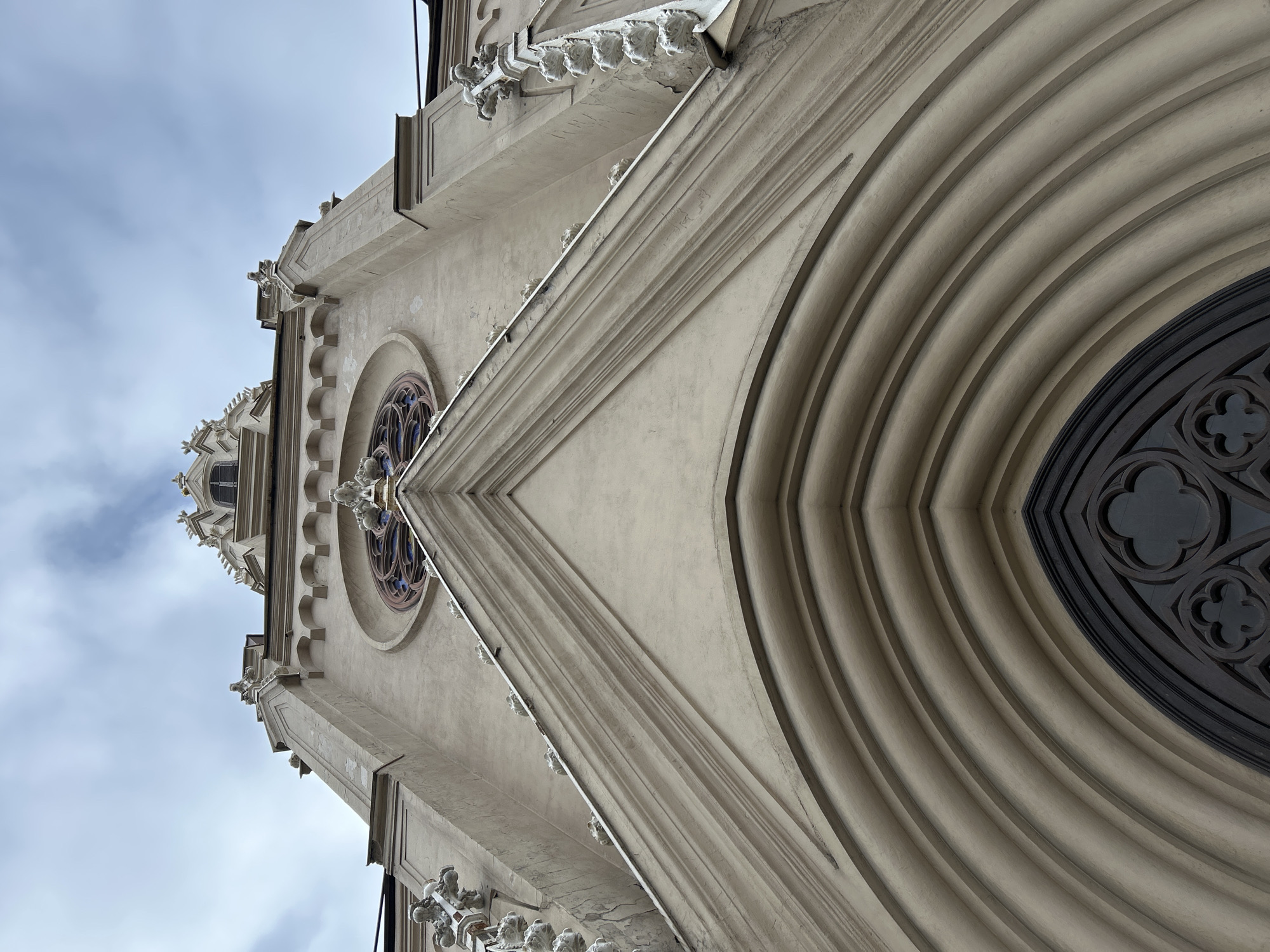 The image showcases a low-angle view of the facade of a building, likely a church or cathedral, identified as the Chesme Church in Saint Petersburg, Russia. The architectural style is Gothic Revival, characterized by pointed arches and decorative stone elements. The facade is light in color, possibly beige or light gray, with intricate details such as moldings, rose windows, and decorative elements resembling gargoyles or pinnacles. A large arched doorway with layered architraves dominates the right side of the frame. The sky is visible in the background, with a mix of clear areas and light clouds. No people are visible in the image.