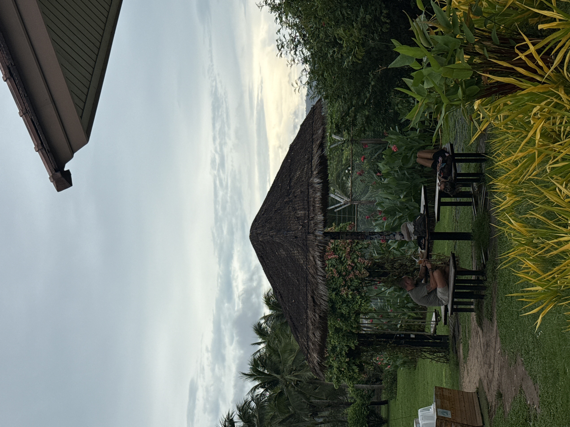 A rotated, medium-shot depicts a thatched hut in บ้านบางรักษ์, Thailand. The sky, which occupies the top half of the photo, is overcast with a hint of sunlight breaking through. A building's edge peeks in from the top left corner. The thatched hut, centrally positioned, is draped with flowering vines and greenery. People, presumably tourists, are seated at a table beneath the hut. The surrounding environment consists of tropical plants and grass.