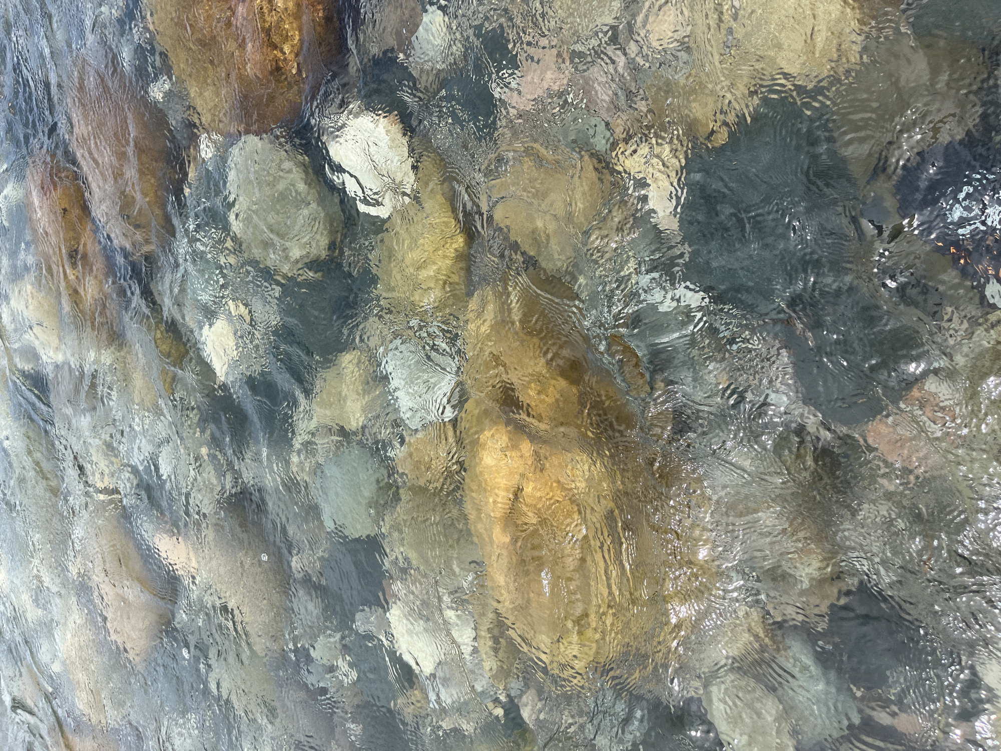 This is a close-up, overhead shot of river rocks beneath clear, flowing water. The rocks vary in size and color, with shades of brown, gray, tan, and white visible. The water creates ripples and distortions on the surface, and the light reflects off the water, creating bright spots and shimmering patterns. The scene appears natural and serene, with no signs of human activity or urban environment. There is no indication of the location being in Majai, India.
