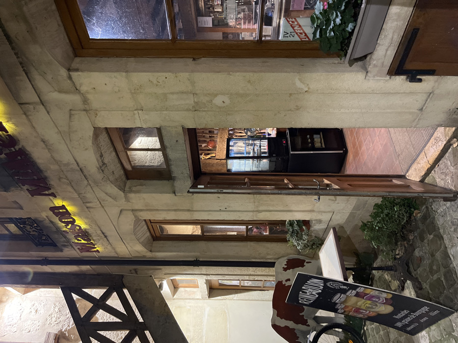 Exterior shot of a restaurant at night. The entrance is open, revealing a refrigerated display case inside. Above the entrance is a window with a wooden frame, and a sign for "Maison Posson" is visible on the building's facade. A small window sits below the entrance. Outside, there's a promotional sign for Reine de Dijon and a decorative bicycle. The building is constructed of stone and the exterior paving consists of cobblestones.