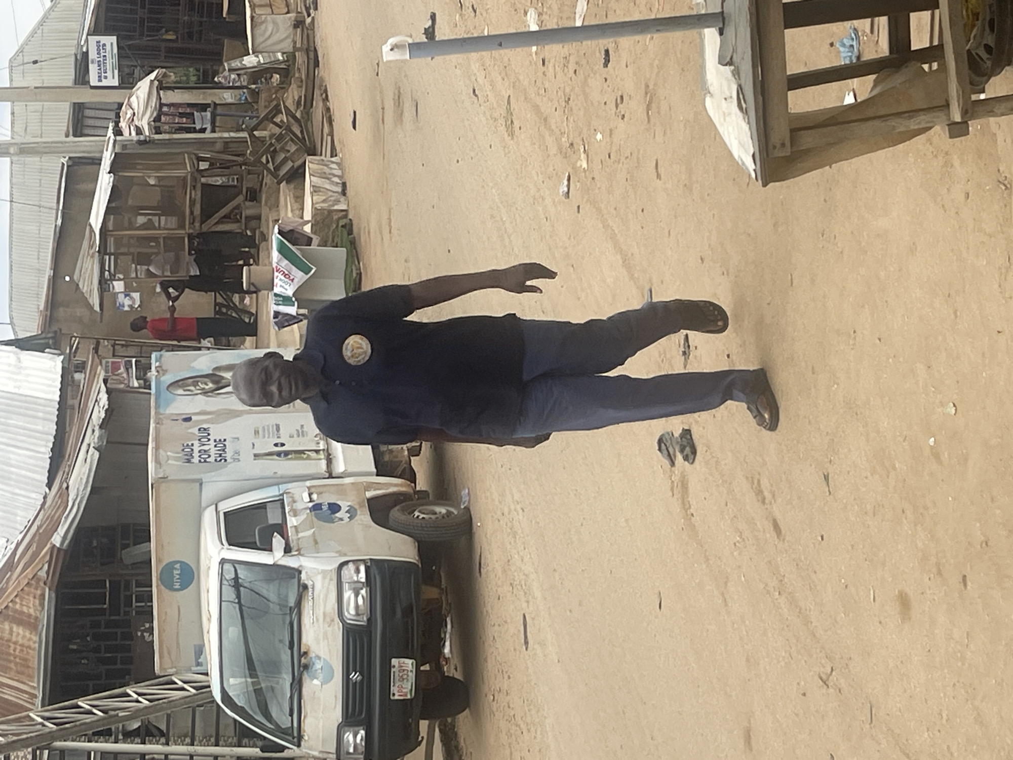 This image shows a street scene in Abuja, Nigeria, with a man in a dark blue uniform standing in the middle of a dusty road. He is facing slightly to his left, with his right arm extended. He appears to be a security guard or someone in a similar role, as indicated by the emblem on his shirt. Behind him, a white truck with "NIVEA" branding is parked. The truck is loaded with large boxes, some of which have images of faces on them, and text that reads "MADE FOR YOUR SHADE".
The background of the image reveals a bustling market or commercial area. There are several market stalls and makeshift structures made of wood and corrugated iron. Some of these stalls display goods, and there are other people in the background, some of whom are partially obscured. A sign for "DREAMS LODGE SUITE LTD" is visible on one of the buildings. The overall impression is of a busy, everyday scene in an outdoor marketplace. The lighting suggests it is daytime, likely mid-morning or afternoon, with bright sunlight casting shadows on the ground. The ground is unpaved and dusty, typical of many roads in this region. The presence of flip-flops on the ground near the man's feet adds a touch of everyday realism to the scene.