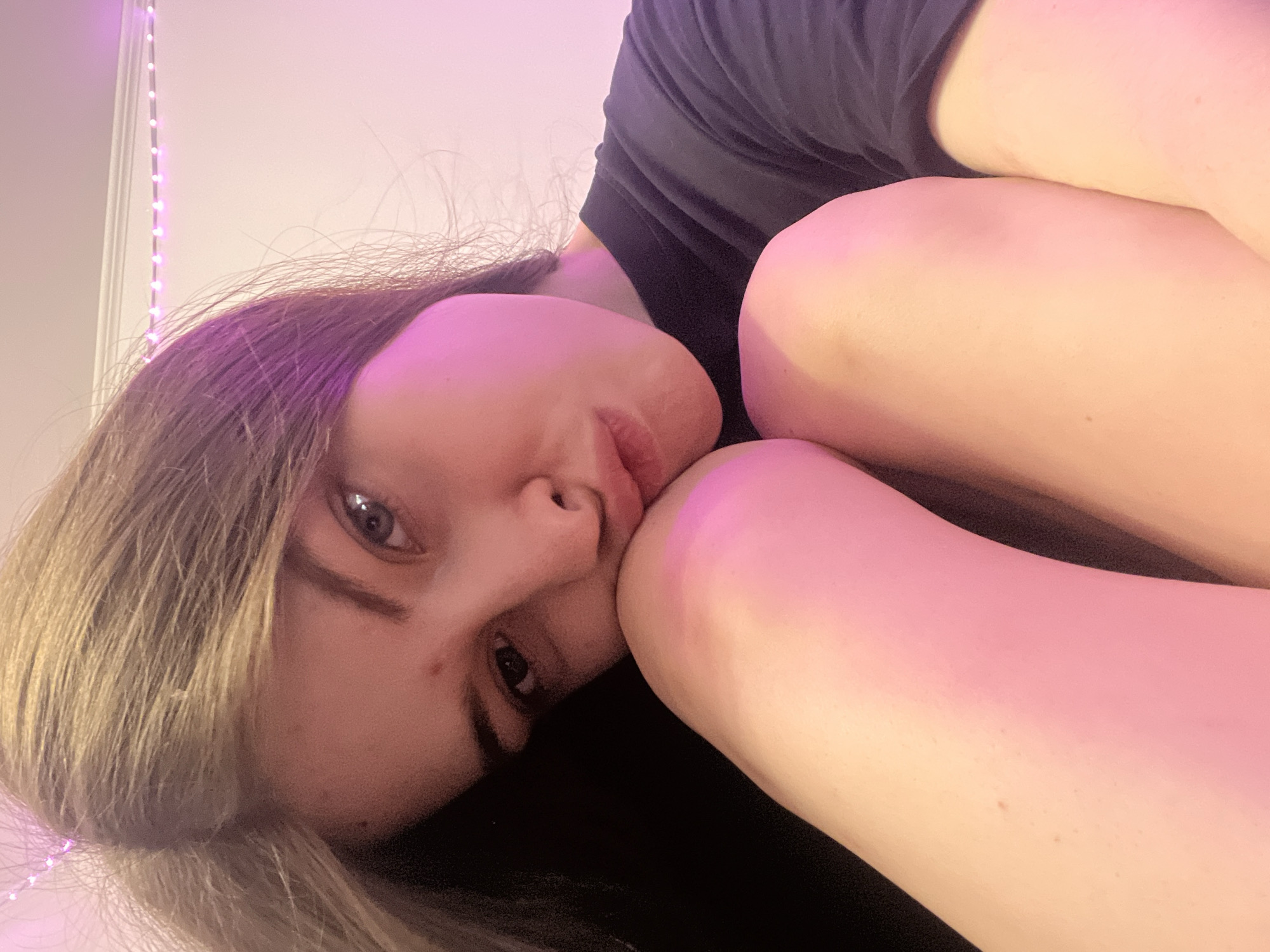 A young woman is lying down, seemingly in a bedroom. Her legs are drawn up towards her chest. She is wearing a dark-colored shirt. The lighting is predominantly pink, possibly from LED lights that are visible in the background. The specific location in Saint Petersburg, Russia, cannot be determined from this image.