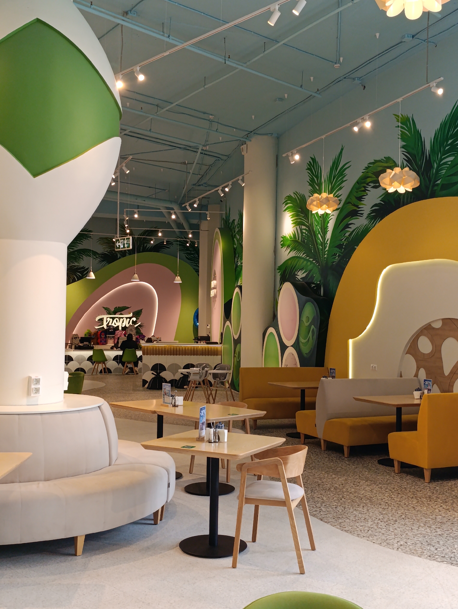 A cafe interior features a tropical-themed design with palm tree murals on the walls. The seating area includes yellow banquettes and chairs, as well as a large, curved beige sofa. Tables are scattered throughout the space, some with condiments and menus. A counter with a neon "Tropic" sign is visible in the background, where a few people are present. The ceiling is painted light blue and has exposed pipes and lighting fixtures. The floor is a light-colored terrazzo. The overall impression is a modern and vibrant cafe.
