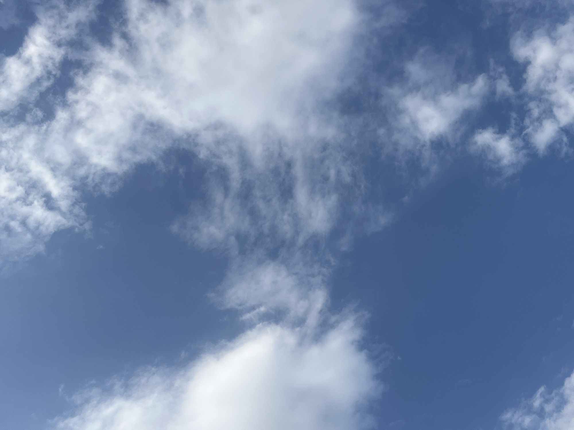 The image depicts a sky with scattered clouds over Batken City, Kyrgyzstan. The clouds are white and fluffy, with some appearing wispy and translucent. The sky is a clear blue, indicating a sunny day. There are no people, animals, or man-made objects visible in the image. The scene is serene and natural, focusing solely on the atmospheric conditions.