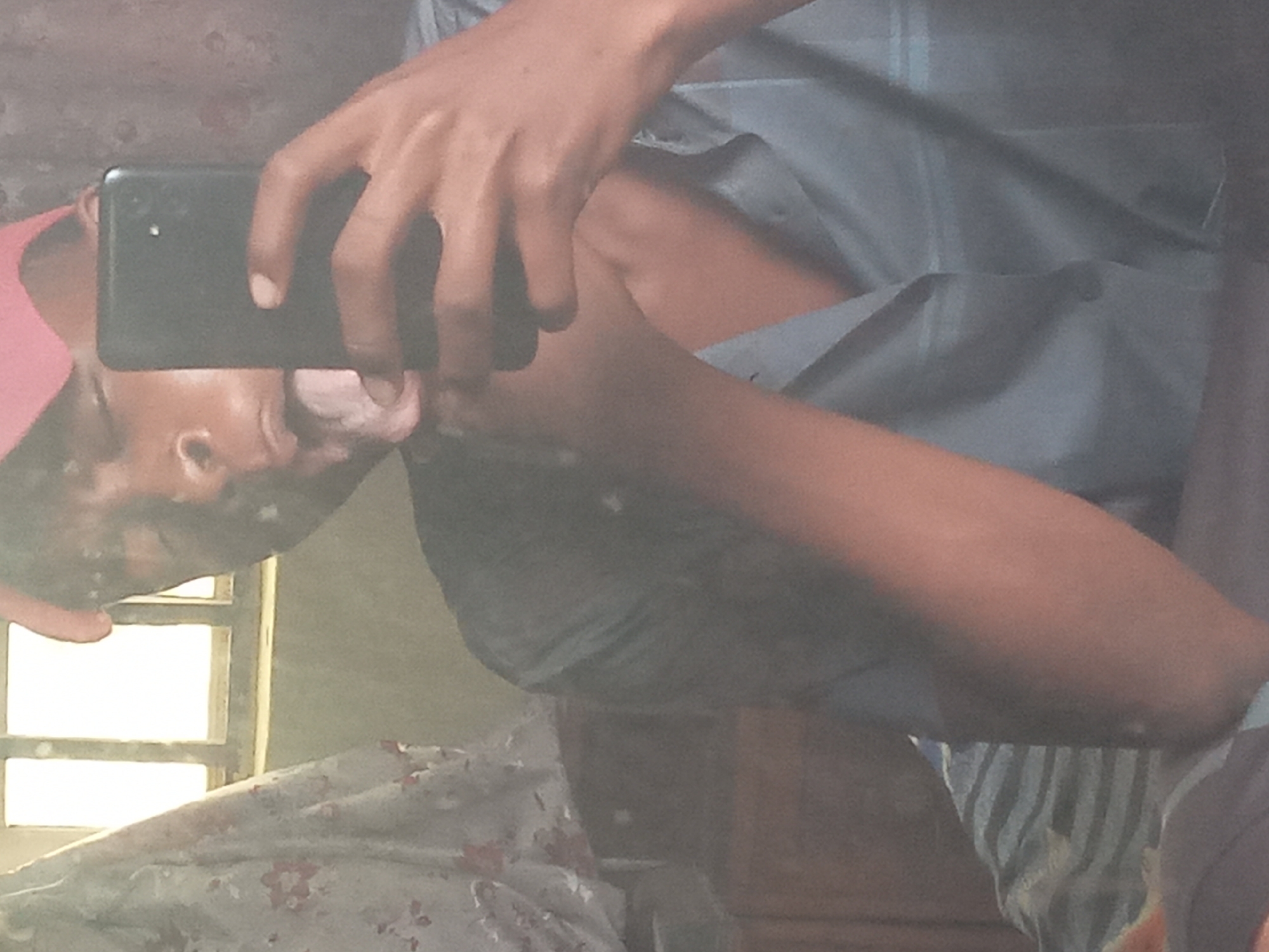 The image shows a person taking a selfie in a mirror. The person is holding a black smartphone. The person appears to be wearing a pink hat and a blue shirt. There is a window with sunlight shining through it in the background. A floral patterned bedsheet is visible at the bottom of the image. There is no information about the location being Onitsha, Nigeria in the image.