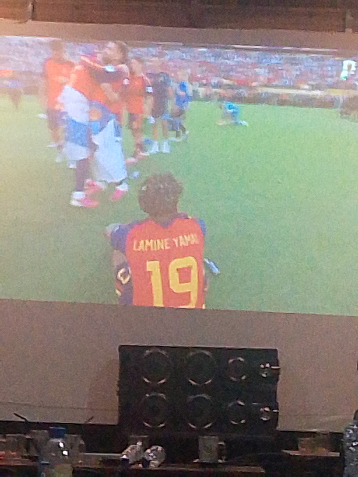 This is a slightly blurry, overexposed, medium shot of a projection of a soccer match on a large screen. The screen displays a green field with many players in red and blue uniforms. A player in the foreground is wearing a red jersey with "LAMINE YAMAL" and the number "19" on the back. The projection is taking place in a dimly lit room with a dark speaker system visible at the bottom of the frame. The top of the image shows a wooden surface, likely a ceiling or overhead structure. The location in Meunasahmee, Indonesia, is not discernible from the image itself.