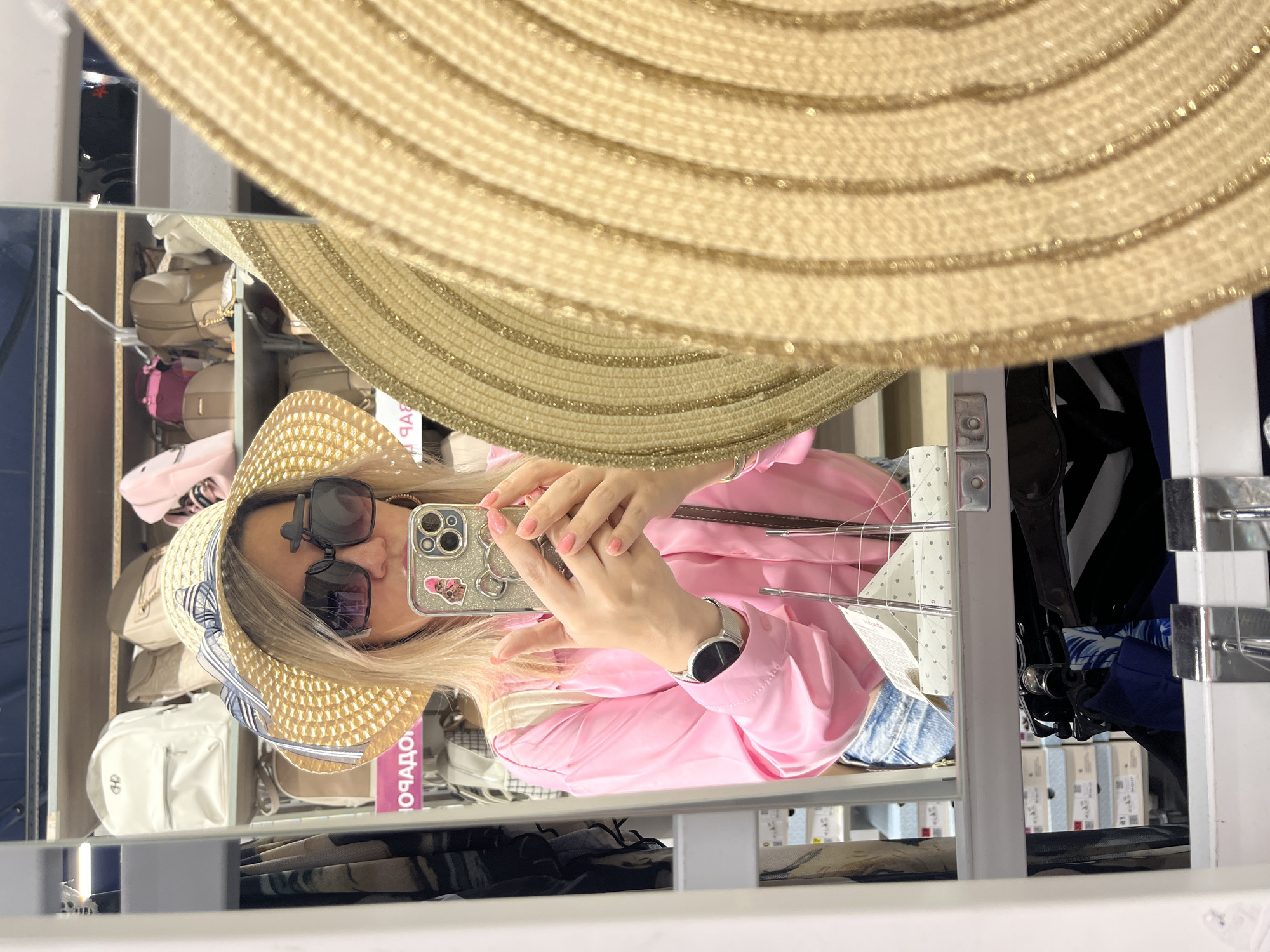 In Sochi, Russia, a young blonde woman in a straw hat and pink shirt takes a selfie in a store mirror. She is holding a smartphone with a Mickey Mouse case and wearing large sunglasses. The background shows shelves stocked with backpacks and other items. A large straw hat is reflected in the foreground of the mirror.