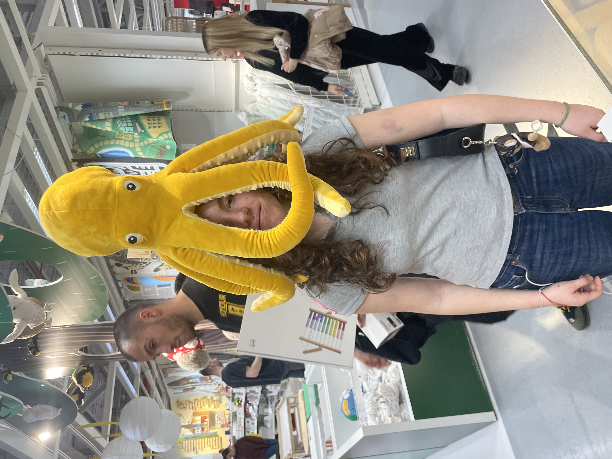 The image, likely taken in a retail setting like IKEA, shows a young woman wearing a yellow plush octopus hat. She has long, curly brown hair and is wearing a gray t-shirt and blue jeans. A black purse hangs over her shoulder. A man in a black t-shirt is visible to her left, holding a white box with a picture of a xylophone on it. In the background are shelves stocked with various items. A woman with long blonde hair wearing black and beige clothing is walking away in the background. The location is not identifiable as Sofia, Bulgaria based solely on the image content.