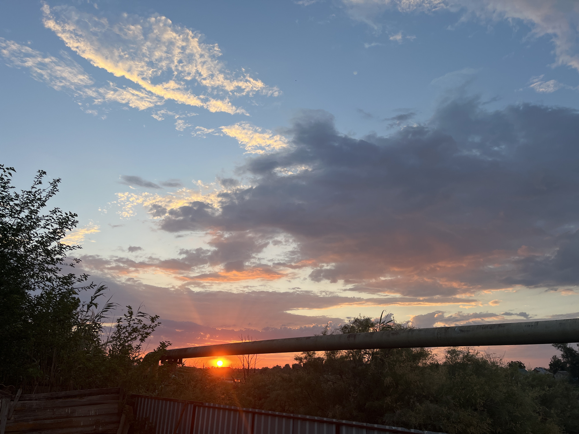 The image depicts a sunset over a rural landscape, likely in or near Krasnyye Barrikady, Russia. The sun, a bright orb of orange, is setting low on the horizon, casting warm light and sunbeams through scattered clouds. The sky transitions from a clear blue at the top to hues of orange, pink, and purple as it nears the horizon, reflecting the sunset. Wispy clouds are illuminated by the sun, while larger, darker clouds dominate the mid-sky. In the foreground, a large, horizontal pipe runs across the frame, obscuring some of the view. To the left and right of the pipe, lush green foliage and trees are visible, partially silhouetted against the bright sky. Below the foliage, a wooden fence and a corrugated metal fence are present, suggesting a boundary or structure. The overall scene is natural and serene, capturing the beauty of a twilight sky in a rustic setting.
