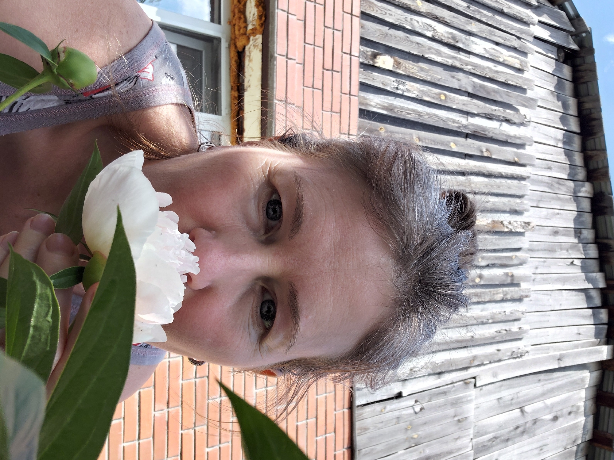 A woman with grey hair pulled back is smelling a white peony in front of a wooden building with a red brick base. The building appears to be a barn or shed with a weathered, slatted wooden exterior. Below the wooden wall is a red tiled roof, and beneath that, exposed insulation is visible around a window. The woman is wearing a grey t-shirt with a pattern. The setting is outdoors, and the lighting suggests it is daytime. The provided context indicates the location is Novosibirsk, Russia, however, there are no visible landmarks or identifiers in the image to confirm this specific location within the city.