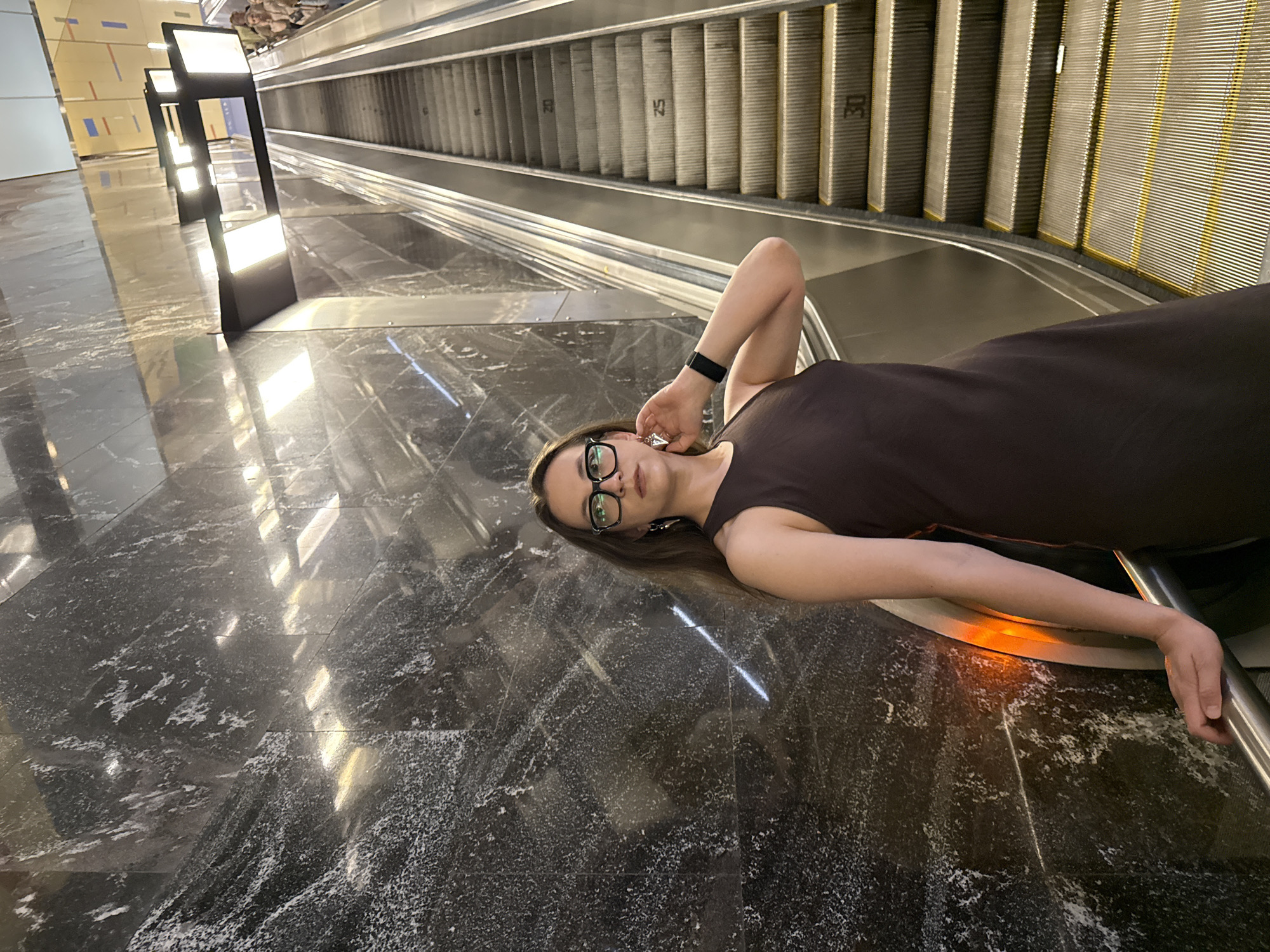 The image captures a young woman lying on a highly reflective, dark marble or granite floor in an indoor public space, likely a metro station or modern building in Moscow, Russia. She is positioned towards the lower right of the frame, looking directly at the camera. She has long brown hair, wears dark-rimmed glasses, a dark brown sleeveless dress, and a black smartwatch on her left wrist. Her left arm is bent with her hand near her head, while her right hand rests near the base of an escalator railing.
Behind her and to the left, a long escalator descends into the background. Its metallic steps are visible, with some steps bearing white numbers, including "25" and "30". The escalator's structure and handrails are clear. To the far left, several sleek, vertical light fixtures with bright panels cast strong, elongated reflections across the polished floor. The floor itself is dark with prominent white veining or speckles, reflecting the artificial lights and elements of the environment. The overall lighting is bright and artificial, typical of an indoor public area, making the time of day indeterminate. The scene conveys a sense of modernity and expansive architecture.