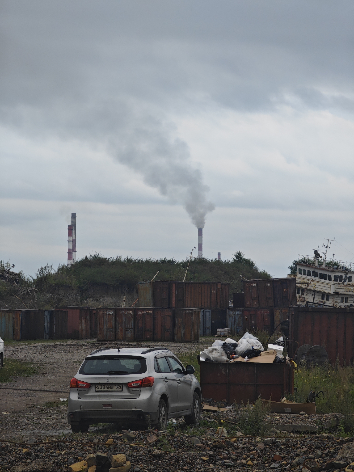 The image depicts an industrial landscape on a cloudy day in Krasnoyarsk, Russia. Two tall, red and white striped smokestacks are visible in the distance, emitting a plume of white smoke, indicating industrial activity. In the foreground, a silver Mitsubishi SUV is parked on a gravel lot. Surrounding the SUV is a collection of rusty shipping containers, some stacked several high, forming a makeshift wall. To the right, a dilapidated white boat, partially submerged or grounded, rests against a shipping container. A large dumpster overflowing with trash is also visible in the middle ground, with plastic bags and cardboard scattered around it. The ground is covered in gravel, rocks, and some sparse vegetation. The overall scene suggests an area of industrial decay or a junkyard, with a stark contrast between the functional smokestacks and the abandoned structures and vehicles.