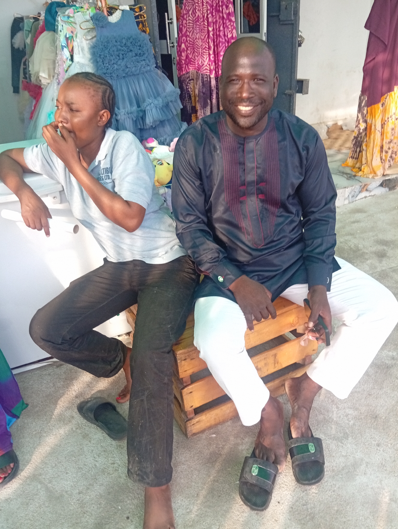 Two people are sitting in front of a store that sells clothing, possibly children's clothing, given the presence of several dresses. The person on the left, a woman with braided hair, is wearing a light grey polo shirt and dark pants. She is leaning on a white surface, possibly a refrigerator or counter, and appears to be looking down and chewing on her fingernail. The person on the right, a man with a shaved head, is wearing a dark long-sleeved shirt with red embroidery and white pants. He is smiling broadly at the camera and holding a phone in his right hand. Both individuals are wearing sandals. The setting appears to be outdoors, with a concrete floor and a doorway in the background. Colorful fabrics hang in the store, suggesting a vibrant marketplace or retail environment. The lighting suggests it is daytime, possibly late afternoon due to the warm tones. The overall atmosphere is relaxed, with the man displaying a cheerful expression. The text "MULTIBIZA RES LTD." is visible on the woman's shirt.