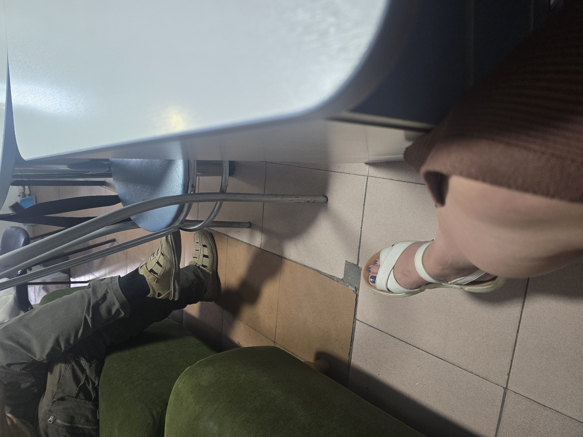 This image depicts an indoor scene. In the foreground, a person's legs are visible from the knee down, wearing a brown knitted skirt and white sandals. Their toenails are painted a dark, shimmery blue. To the left, another person is seated on a green couch, wearing dark pants, black socks, and tan open-toed sandals. Behind them, a chair with a blue seat and metal legs is partially visible, alongside a table with a white surface. The floor is tiled in a neutral tone. The overall setting suggests a casual indoor environment, possibly a waiting area or a common space. There is no information in the image to indicate the location is the city of Ulan-Ude, Russia.