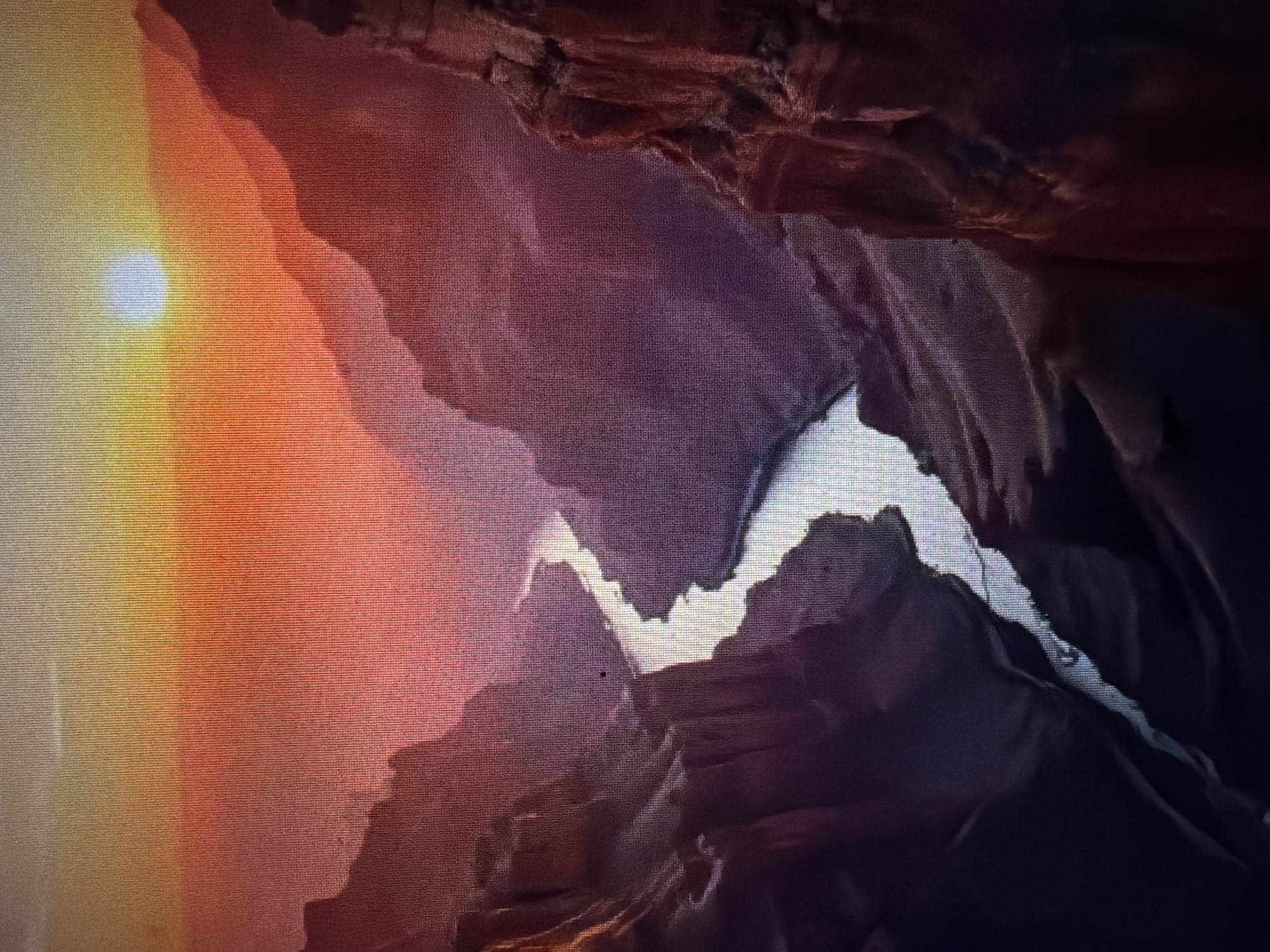 The image is a high-angle, wide shot of a canyon with a river flowing through it, illuminated by the setting sun. The canyon walls are steep and rugged, with layers of rock visible. The sky is a gradient of orange, yellow, and pink, with the sun appearing as a bright white orb near the horizon. The river reflects the colors of the sky, appearing as a silvery ribbon winding through the canyon. The scene is peaceful and majestic. The provided crops of the image show close-ups of the sky and canyon walls, and the river. There is no indication of the city of Baan Mae Nam, Thailand, or any specific location in the image.