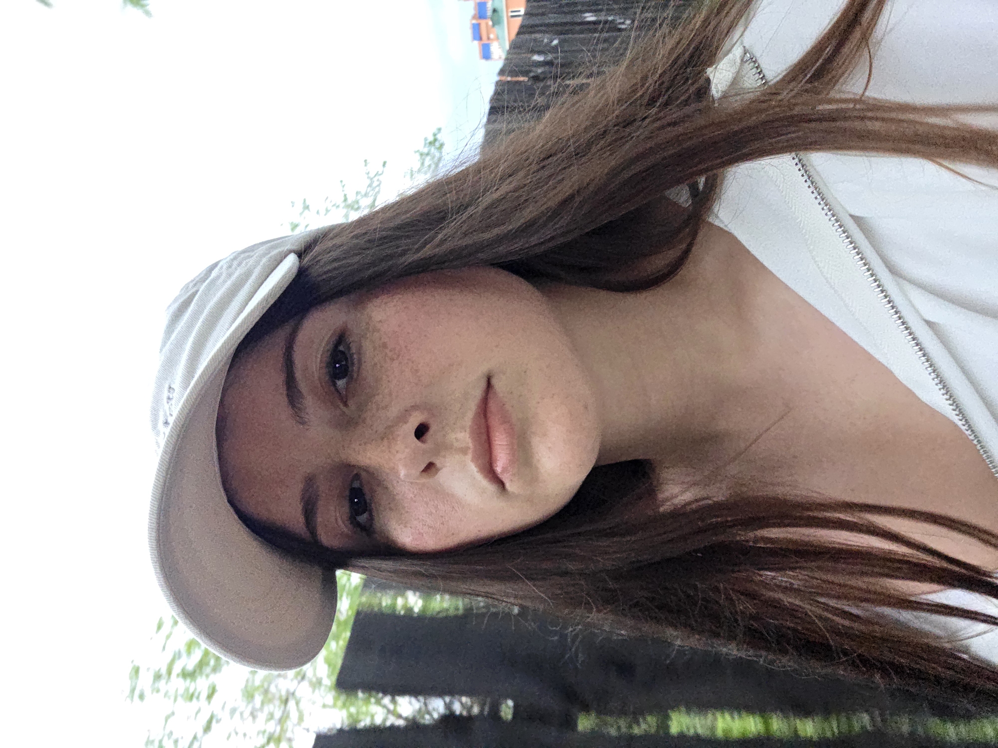 This media file is a close-up, rotated selfie of a young woman outdoors. She is wearing a light-colored baseball cap and a white zippered jacket. Her long, dark brown hair is flowing around her face and shoulder. She has fair skin with freckles and is looking towards the camera with a neutral expression.
The background is out of focus, but appears to be an outdoor setting with some greenery and what looks like a wooden fence or structure. Above the fence, there are some colorful structures that could be part of a building or some sort of decorative element. The lighting suggests it is daytime, possibly a bright day with overcast skies given the whiteness of the background. The overall impression is casual and informal.