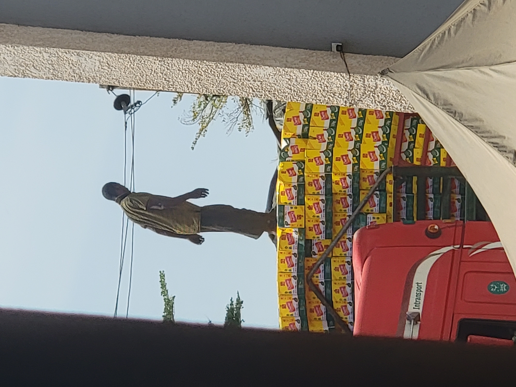 The image, taken in Maiburiji, Nigeria, shows a mannequin suspended in the air by wires against a clear, light blue sky. The mannequin wears a tan, short-sleeved shirt and dark shorts. Behind the mannequin, a stack of yellow and green packages is visible, partially obscuring a red truck cab with the word "Intransport" displayed on its side. Part of a grey umbrella is visible in the upper right corner of the image. The scene is framed by a light-colored textured overhang at the top and a dark, out-of-focus area in the foreground.