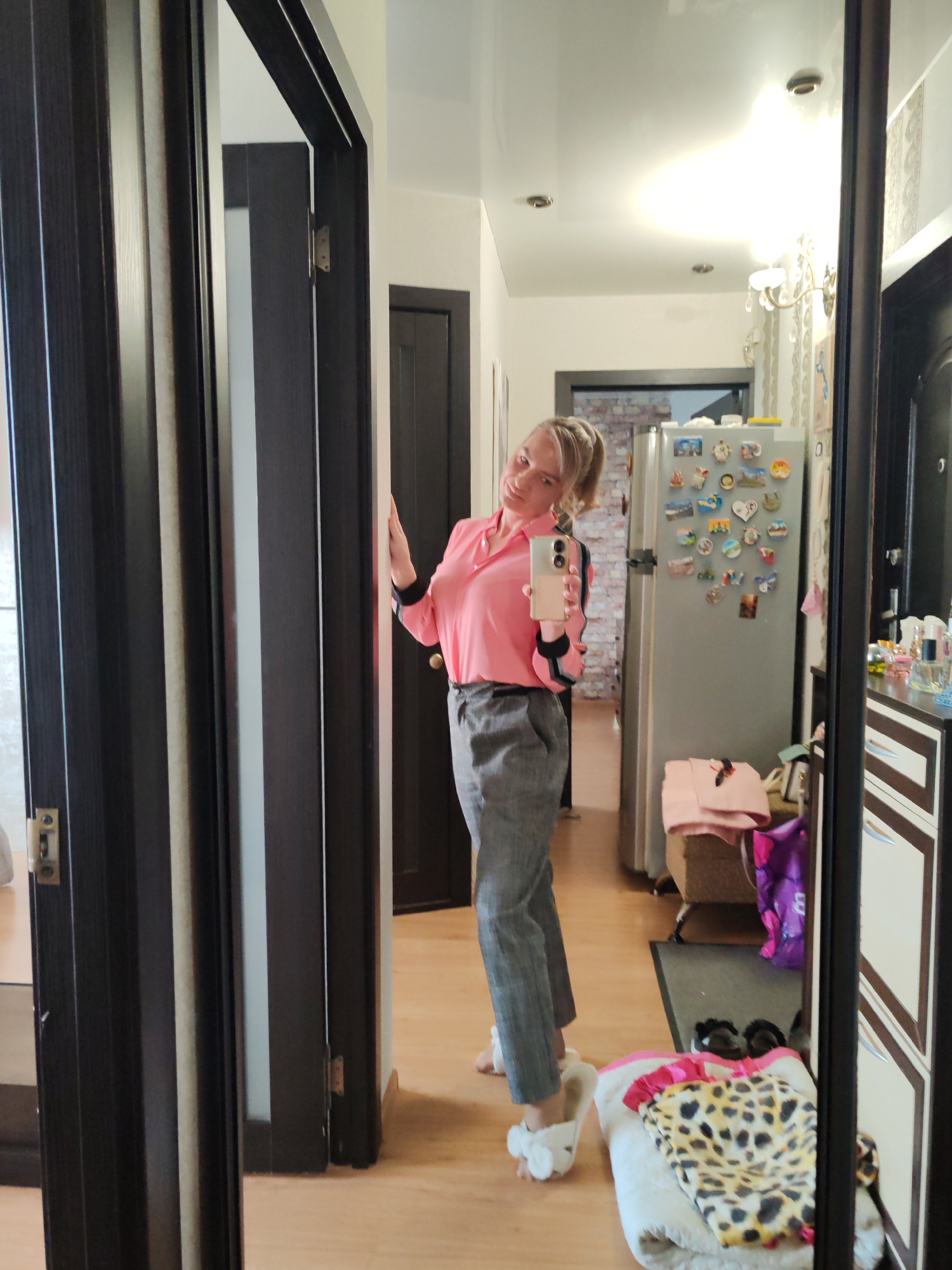 A woman is taking a mirror selfie in a hallway. She has blonde hair pulled back in a ponytail and is wearing a pink long-sleeved shirt with black and white stripes on the cuffs and collar. She is also wearing grey checkered pants and white bunny slippers. The hallway appears to be in a residential apartment.
Visible objects include a refrigerator covered in magnets, a dresser with several drawers, a basket of laundry, and a large mirror. The floor is made of light-colored wood. The overall lighting suggests it is daytime, and there are no windows visible to indicate the weather. The woman's pose and smile suggest a casual and lighthearted mood.