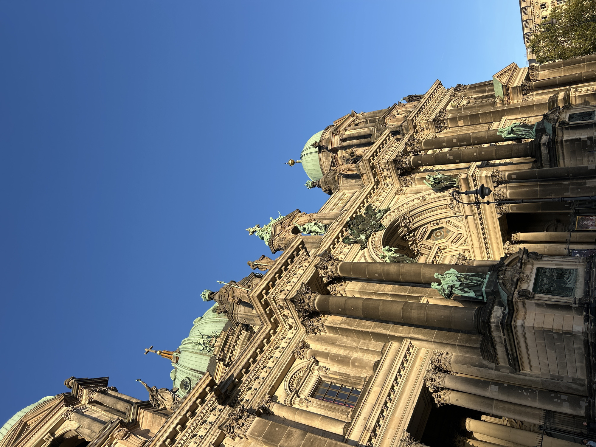 The image shows the Berlin Cathedral (Berliner Dom), a magnificent Protestant cathedral located on the Museum Island in the Mitte district of Berlin, Germany. The photograph is taken from a low angle, looking up at the imposing facade of the cathedral. The building is constructed of stone and features ornate architectural details, including columns, sculptures, and domes. The main dome, covered in verdigris, is prominently visible against a clear blue sky. Several statues, some appearing to be made of bronze, are integrated into the architecture, depicting human figures. Sunlight casts strong shadows on the building, highlighting its textured surfaces and the depth of its features. The scene is serene, with no people clearly visible in the immediate foreground, suggesting a quiet moment of observation. The overall impression is one of grandeur and historical significance.