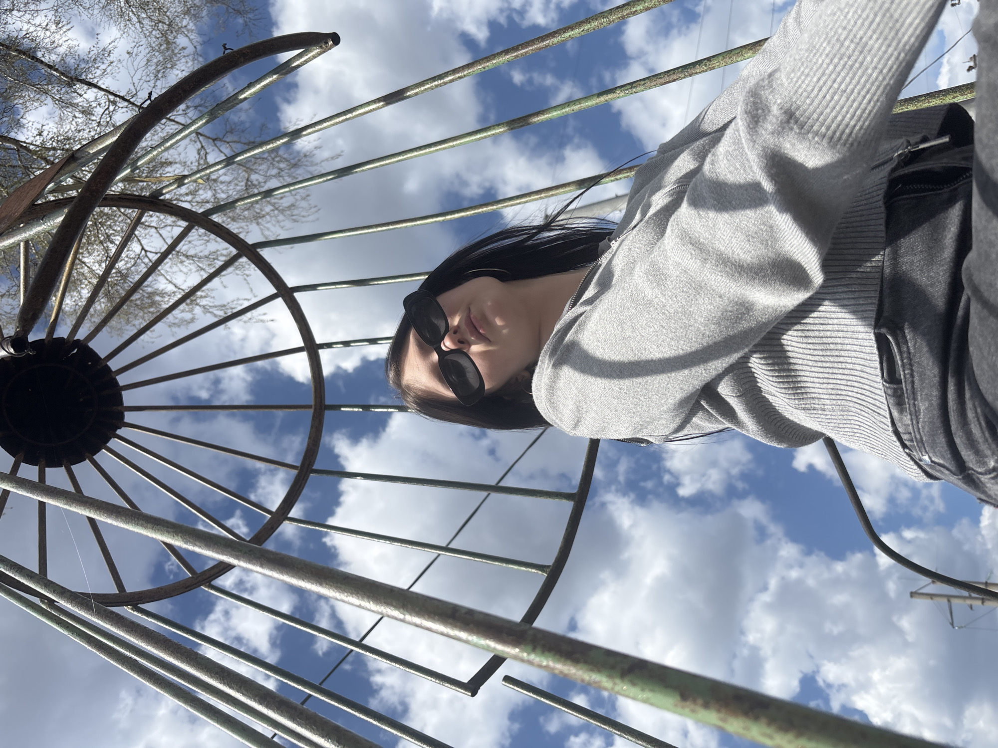 A woman with long black hair and sunglasses, wearing a gray sweater, is positioned under a rusty, wheel-like metal structure. The woman is on the right side of the frame, angled slightly downwards. The metal structure, resembling a portion of a wheel or perhaps an abstract art piece, consists of a circular hub with multiple radiating bars against a backdrop of blue sky dotted with fluffy white clouds. The location is presumably Фоки, Russia, based on the prompt.