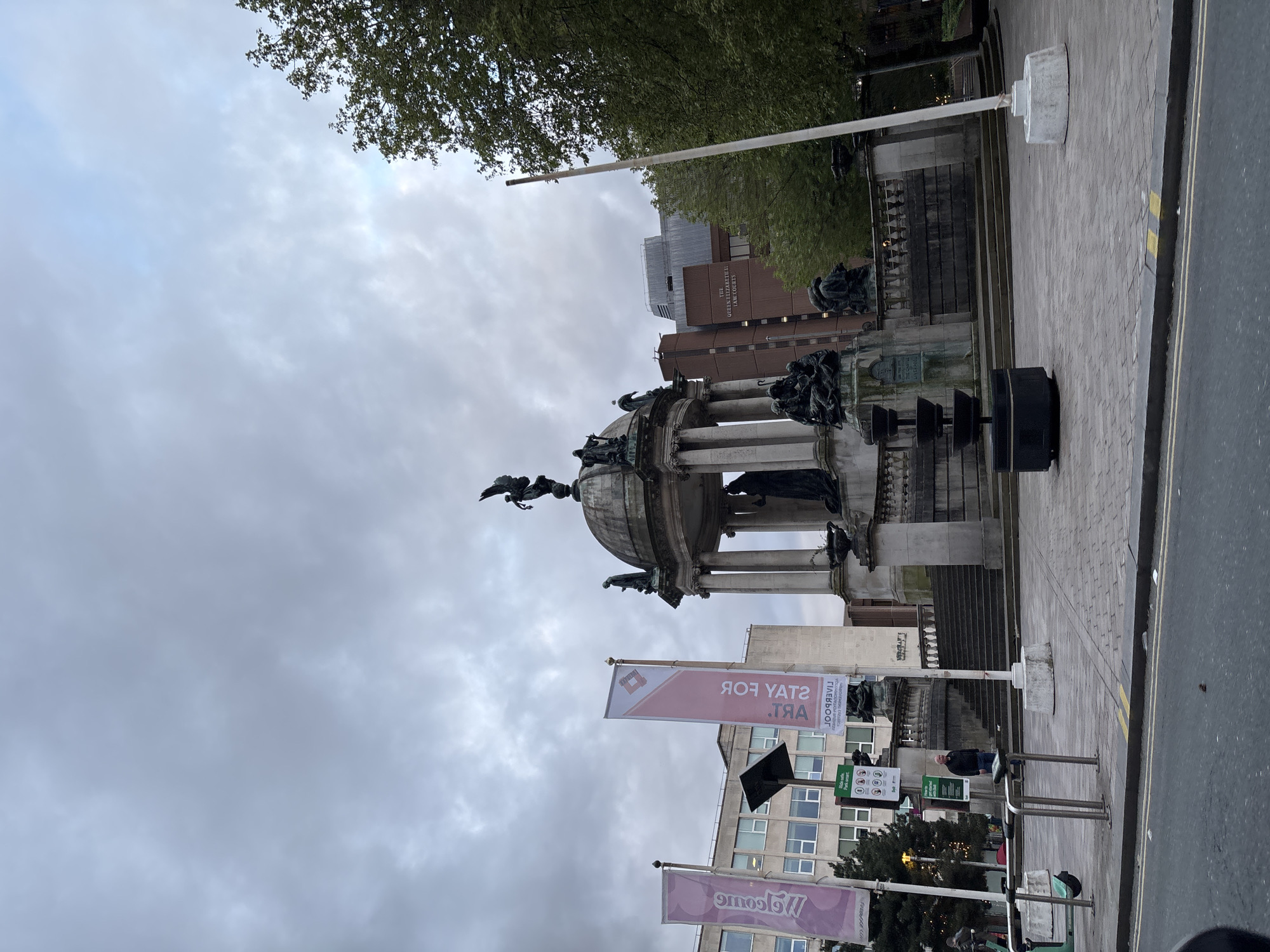 This image depicts the Queen Victoria Monument in Liverpool, United Kingdom. The monument is a large, ornate structure made of stone with columns and sculptures. A winged figure stands atop the dome.
The monument is situated on a paved area alongside a road. The sky is overcast with grey clouds.
There are several banners that say "Stay for Art." A building with windows is visible in the background. The architecture of nearby structures indicates a city environment.