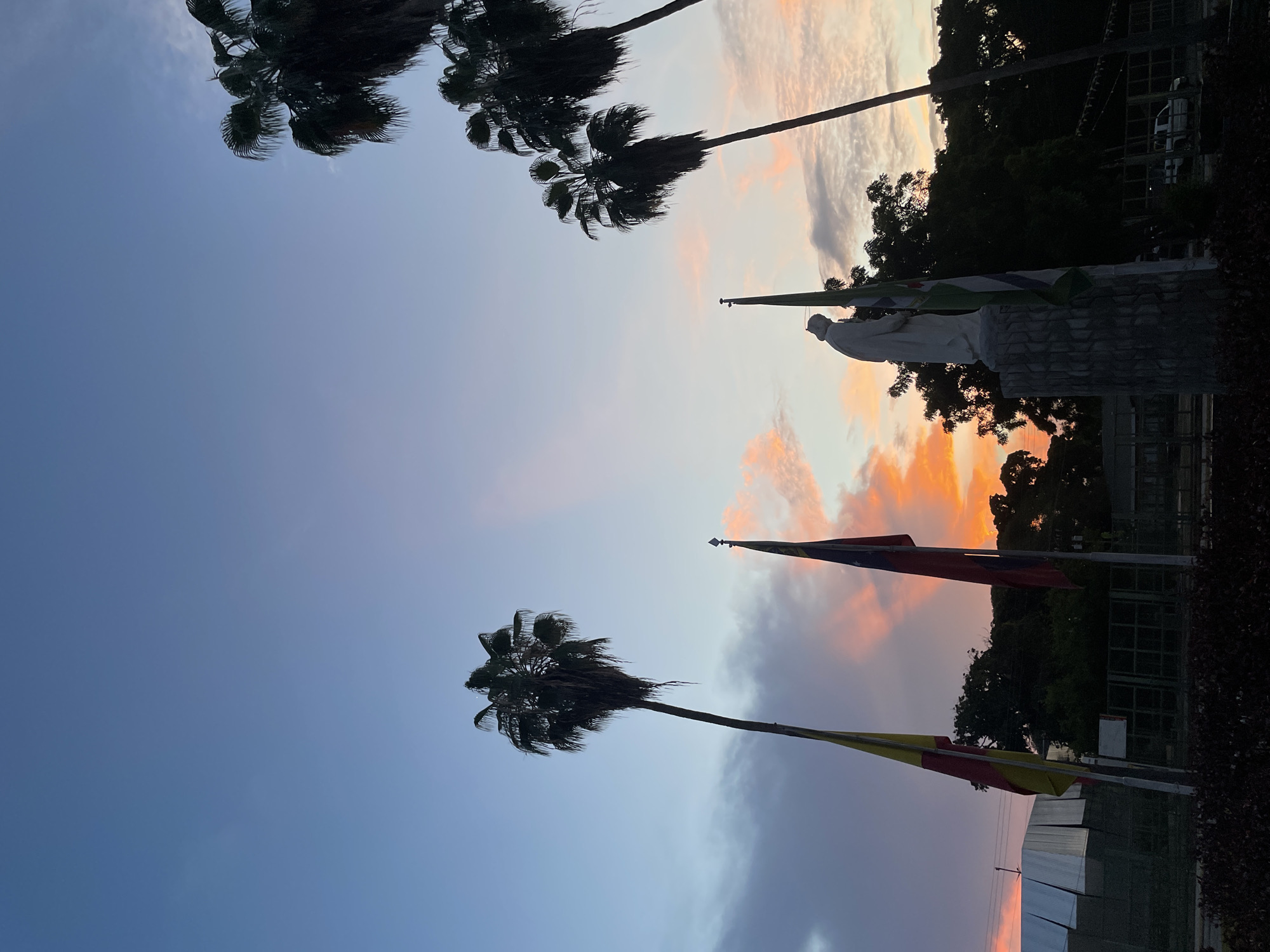The image depicts a sunset scene in Maracay, Venezuela, likely at a monument or public square. In the foreground, a statue of a robed figure stands on a stone pedestal, flanked by flags. Two palm trees are prominent in the frame, silhouetted against a sky painted with hues of orange, pink, and blue, indicative of twilight. The setting sun casts a warm glow, illuminating the clouds and creating shafts of light. The background shows more palm trees, dense foliage, a fence, and a modern building with a metallic sheen on the left. A white vehicle is visible in the distance. The overall atmosphere is serene and picturesque.