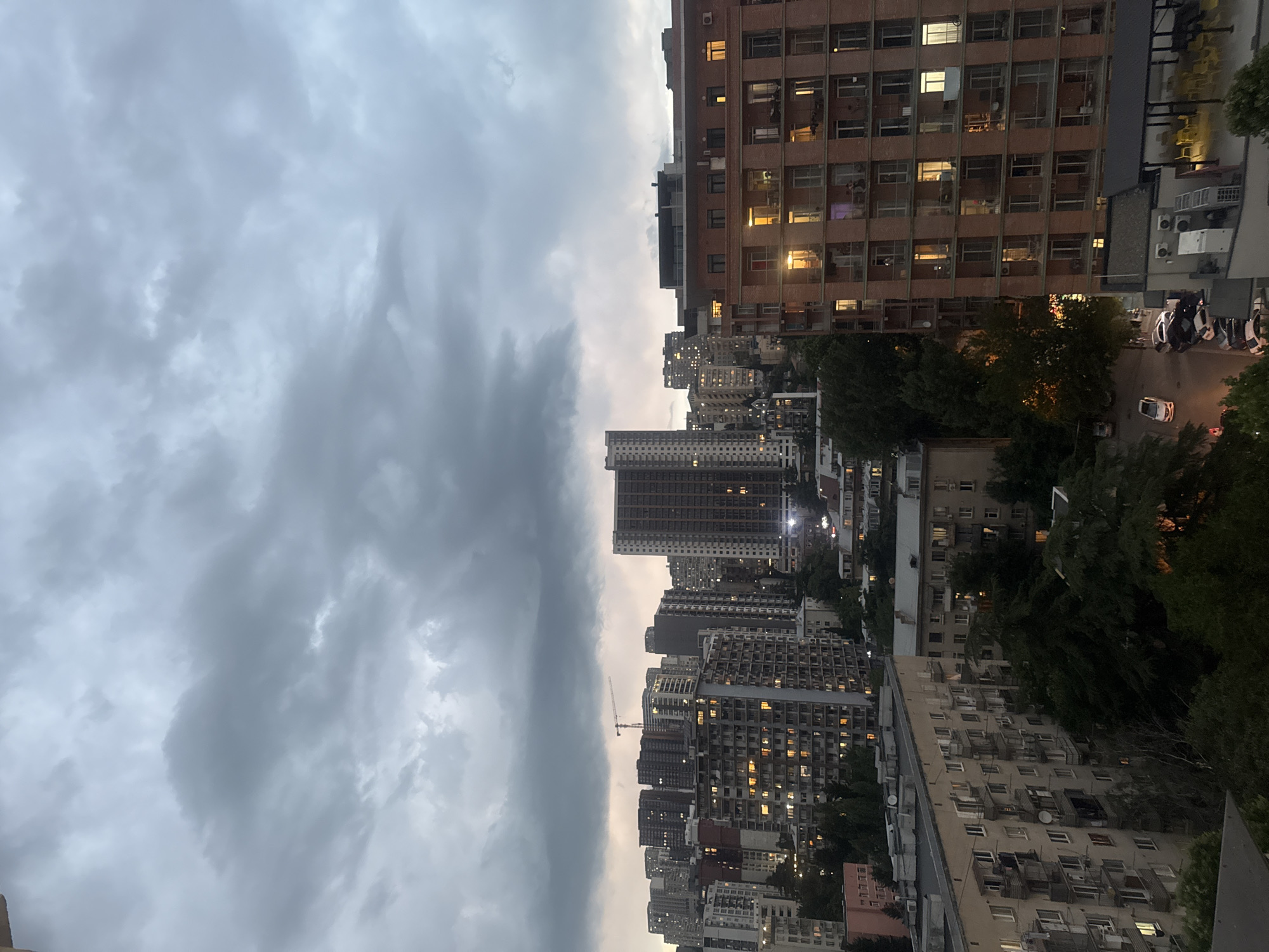 The image captures an urban scene at dusk, likely in Tbilisi, Georgia. The foreground is dominated by a dramatic, cloudy sky, suggesting overcast weather or an impending storm. The sky takes up roughly the left half of the frame, with swirling grey clouds creating a sense of depth and atmosphere.
The right half of the image showcases a dense collection of apartment buildings. These buildings vary in height and architectural style, with many featuring multiple windows, some of which are illuminated, indicating that people are inside their homes. One prominent building on the right is made of reddish-brown brick, with a grid-like pattern of windows. Further back, a cluster of taller, more modern buildings rises, their lights twinkling against the dimming sky. In the mid-ground, trees interspersed among the buildings add a touch of greenery to the urban landscape.
A street is visible at the bottom right, with several parked cars and one white car in motion. This suggests ongoing activity in the city despite the approaching darkness. A construction crane is visible in the distance, hinting at ongoing development. The overall mood is somewhat somber due to the cloudy sky and approaching night, yet the illuminated windows offer a sense of warmth and human presence. There is no visible text in the image.