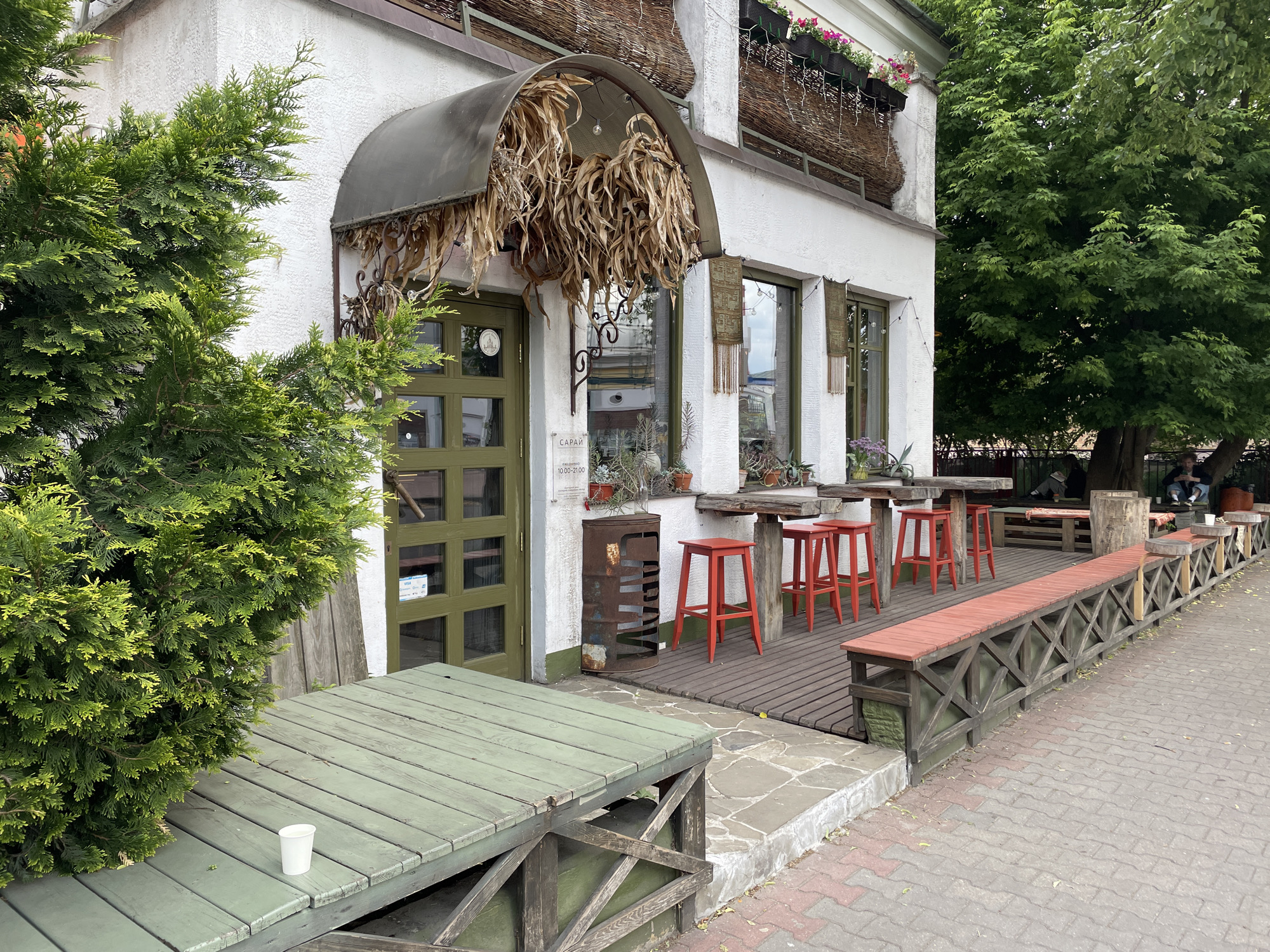 The image depicts the exterior of a building, likely a cafe or restaurant, located in Brest, Belarus. The building is white with green trim around the door and windows. The entrance has a unique awning decorated with dried corn stalks. There is outdoor seating, including red stools at a wooden bar along the building's facade, and green wooden tables and benches nearby. A large green bush stands to the left of the entrance. The walkway is paved with red brick. People can be seen seated further down the walkway in the background.