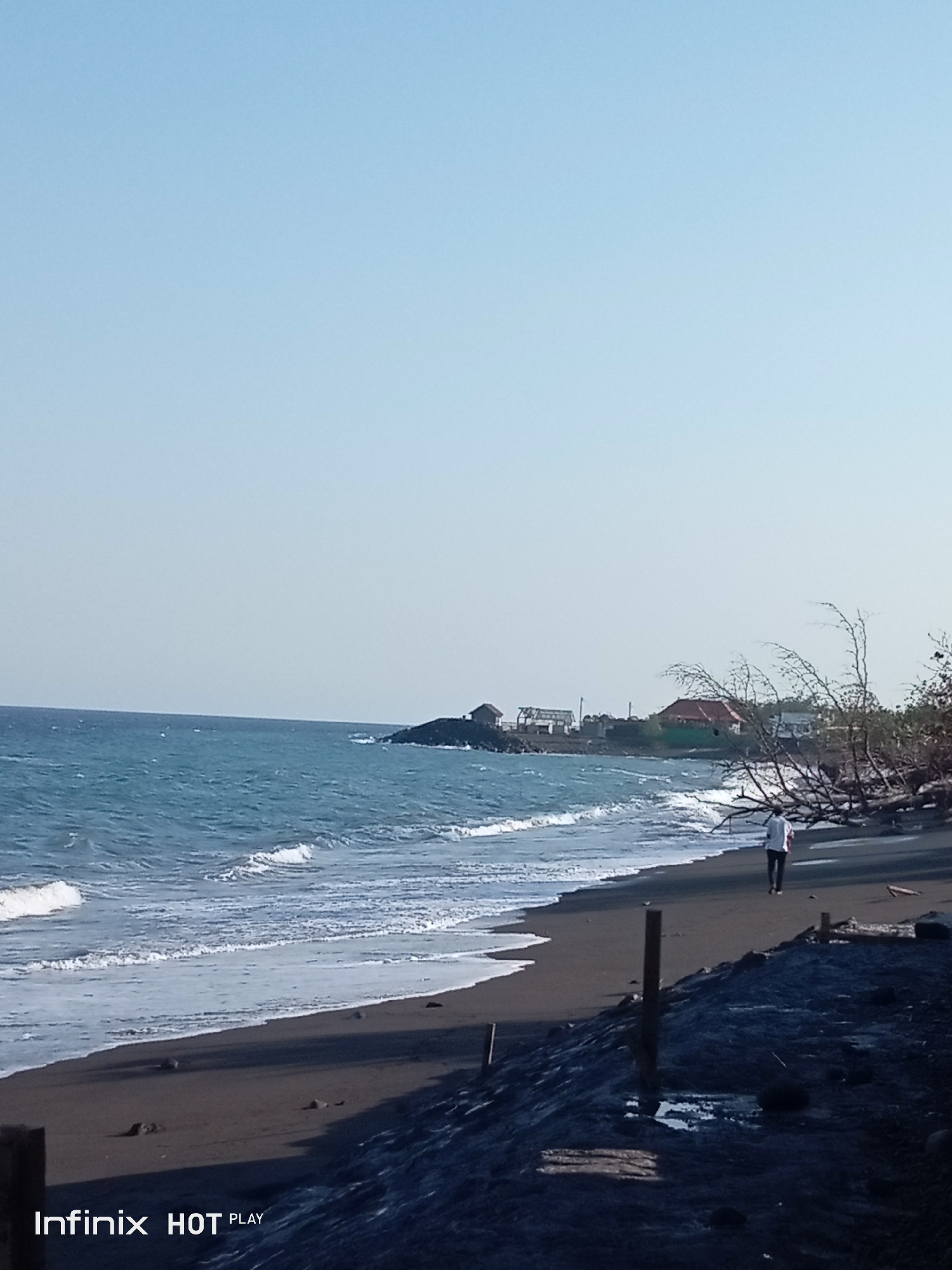 The image depicts a serene coastal scene on a clear day, likely in Dawan, Indonesia, as suggested by the watermark "Infinix HOT PLAY". The foreground is dominated by a dark, sandy beach with some scattered debris and a few wooden posts. A person in a white shirt and dark pants is walking away from the camera along the shoreline, towards the ocean. The middle ground features the ocean, with gentle waves lapping against the shore and a distinct white foam line. In the background, a rocky outcrop juts into the sea, and on this outcrop, there are a few small buildings, one with a red-tiled roof. Further along the coast, a line of trees, some appearing dead and bare, adds to the natural landscape. The sky is a pale, clear blue, indicating a bright and possibly warm day.