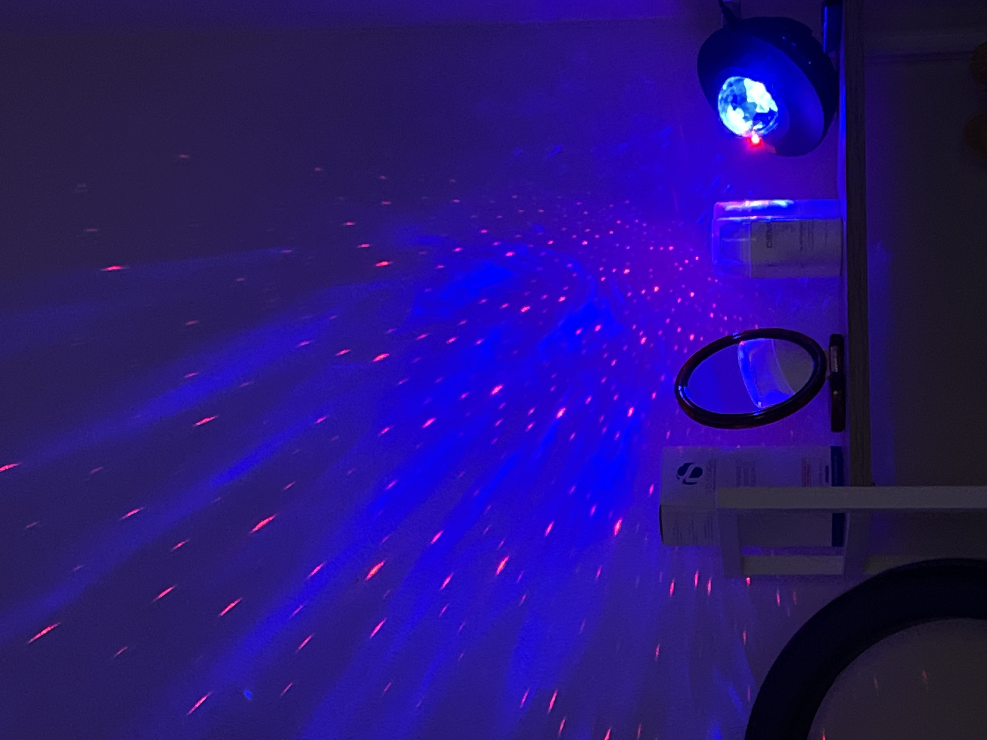 The image depicts an indoor scene characterized by striking artificial lighting, suggesting a darkened room or nighttime setting. A disco ball light projector, mounted on the upper right, is the primary light source, casting a vibrant blue glow across the wall on the left. This blue light is interspersed with numerous red, laser-like dots and streaks that create a dynamic, swirling pattern across the wall, evoking a party or sensory room ambiance.
On the right side of the frame, a light-colored shelf or cabinet structure is visible. On an upper shelf, there is a white rectangular box featuring a distinctive blue, stylized logo that appears to be a linked shape or a letter, possibly a 'C'. Faint text on this box includes the partial word "DERM". Below this shelf, a round mirror with a black frame is positioned. A lower shelf is also present, holding another similar white box with the same blue logo and less discernible text. A portion of another large, black-framed round object is visible at the very bottom right of the image. There are no people visible in the scene. The overall impression is one of lively, colorful illumination in Budva, Montenegro.