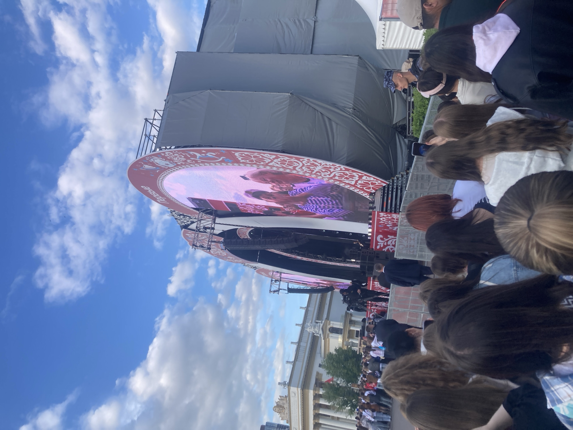 This image depicts an outdoor public event in Moscow, Russia, under a clear blue sky dotted with white clouds. A large crowd of people is gathered, mostly seen from behind, facing a substantial stage. The stage features a prominent circular LED screen displaying a close-up image or video of two individuals, possibly performers or presenters. The screen is adorned with a decorative red and white border featuring traditional patterns and the Cyrillic text "ПЕРВЫЕ" (meaning "FIRST" or "THE FIRST ONES").
The stage structure is robust, with visible scaffolding, dark coverings, and sound equipment (speakers) mounted on the sides. In the foreground, the heads of numerous spectators are visible, with some individuals holding up smartphones, likely to record or photograph the event. Behind the stage, classical-style buildings with columns are partially visible, hinting at a grand public square or exhibition grounds. The bright natural light indicates it is daytime. The scene suggests a concert, festival, or public gathering.