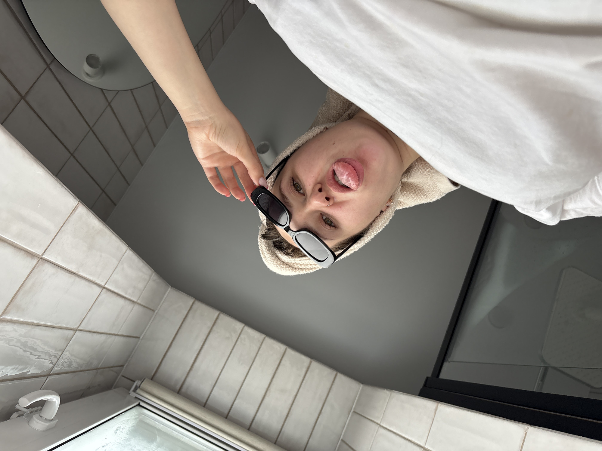 A person with a towel-wrapped head, wearing sunglasses, is sticking their tongue out in a bathroom. The bathroom has white tiles on the walls and a window in the corner. A sink with a mirror is visible. There is no indication of Izhevsk, Russia in the image.