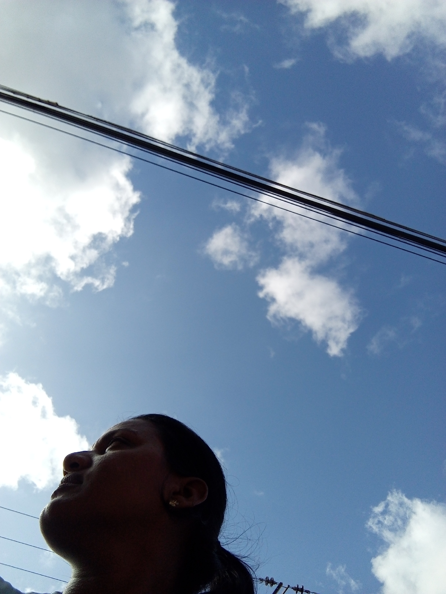 A low-angle, medium shot looking upward shows a person’s face and neck in the lower left corner, power lines running across the top of the image, and a blue sky with white clouds in the background.
The person is facing upward, with their nose, lips, and chin visible in profile. The person's skin tone appears dark and may be partially obscured by shadow. An earring is visible in their ear.
Several black power lines run across the top of the image.
The sky is blue with scattered white clouds.