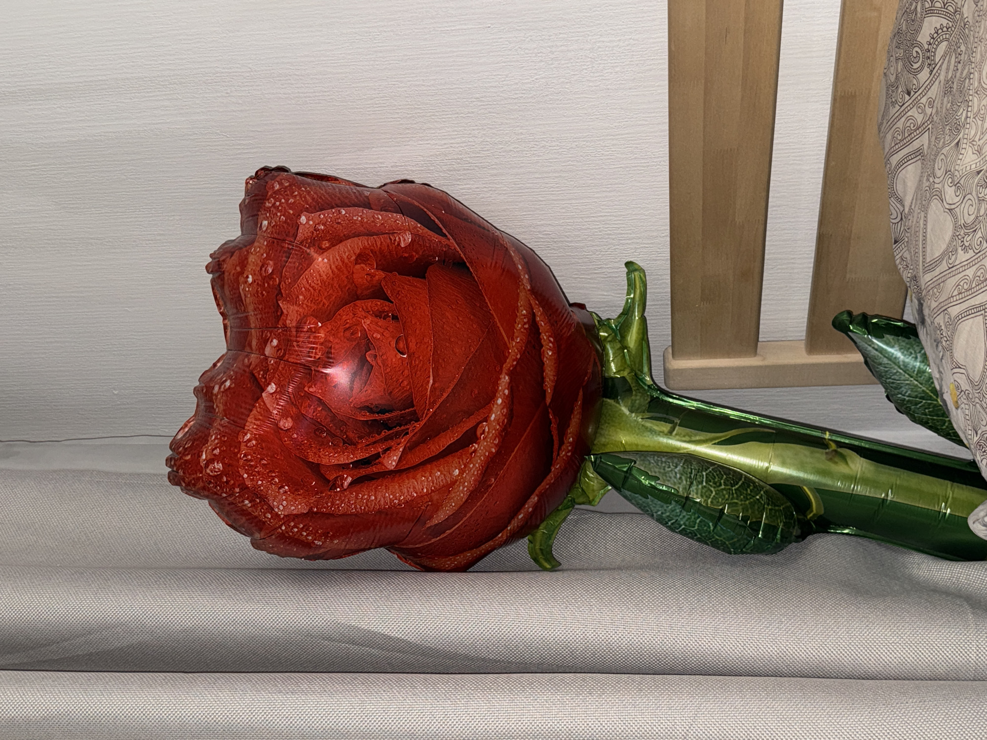 The image displays a close-up, horizontal shot of a vibrant red rose-shaped foil balloon resting indoors. The balloon is highly detailed, featuring printed elements that mimic the texture of rose petals, complete with simulated water droplets for a dewy effect. A shiny green foil stem and leaves extend from the rose, also patterned with vein-like details.
The balloon lies on a light grey fabric surface, possibly a bedspread or sofa cover, which shows a subtle woven texture and two soft, horizontal folds. In the background, a light-colored, textured wall is visible across the left and central portions. To the right, a segment of a light brown wooden structure, composed of vertical slats, can be seen, likely part of a headboard or furniture. Adjacent to the wooden piece, a section of a patterned fabric, possibly a pillowcase or blanket with an intricate, darker design on a lighter background, is partially visible.
The scene is illuminated by artificial indoor lighting, creating a gentle highlight on the reflective surface of the balloon. There is no discernible activity taking place, and no people are present. No text is visible anywhere in the image. While the location context is given as Saint Petersburg, Russia, there are no specific visual cues within the photograph to confirm this.