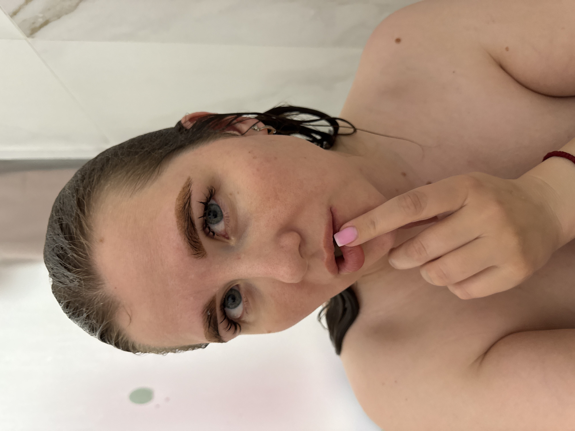 An image captures a young woman from the chest up, positioned as if lying down or reclining, with her head slightly tilted towards the viewer. Her dark hair is wet and slicked back, indicating she has recently bathed or is in a water-related setting. Her striking blue eyes gaze directly into the camera, framed by defined eyebrows and long, dark eyelashes. Her lips are slightly parted, and her left index finger, painted with light pink nail polish, gently touches her lower lip. Her skin is fair, with several small moles visible on her face and bare shoulders. A simple red string or bracelet is visible on her right wrist. The background consists of bright, plain white surfaces, likely tiled walls of a bathroom, with a faint greenish spot visible in the lower left, possibly an object or reflection in bathwater. The lighting is soft and even, highlighting her features. The scene suggests a private moment, possibly contemplative or posed, within a bathroom setting in Ферма, Russia. No text is visible in the image.