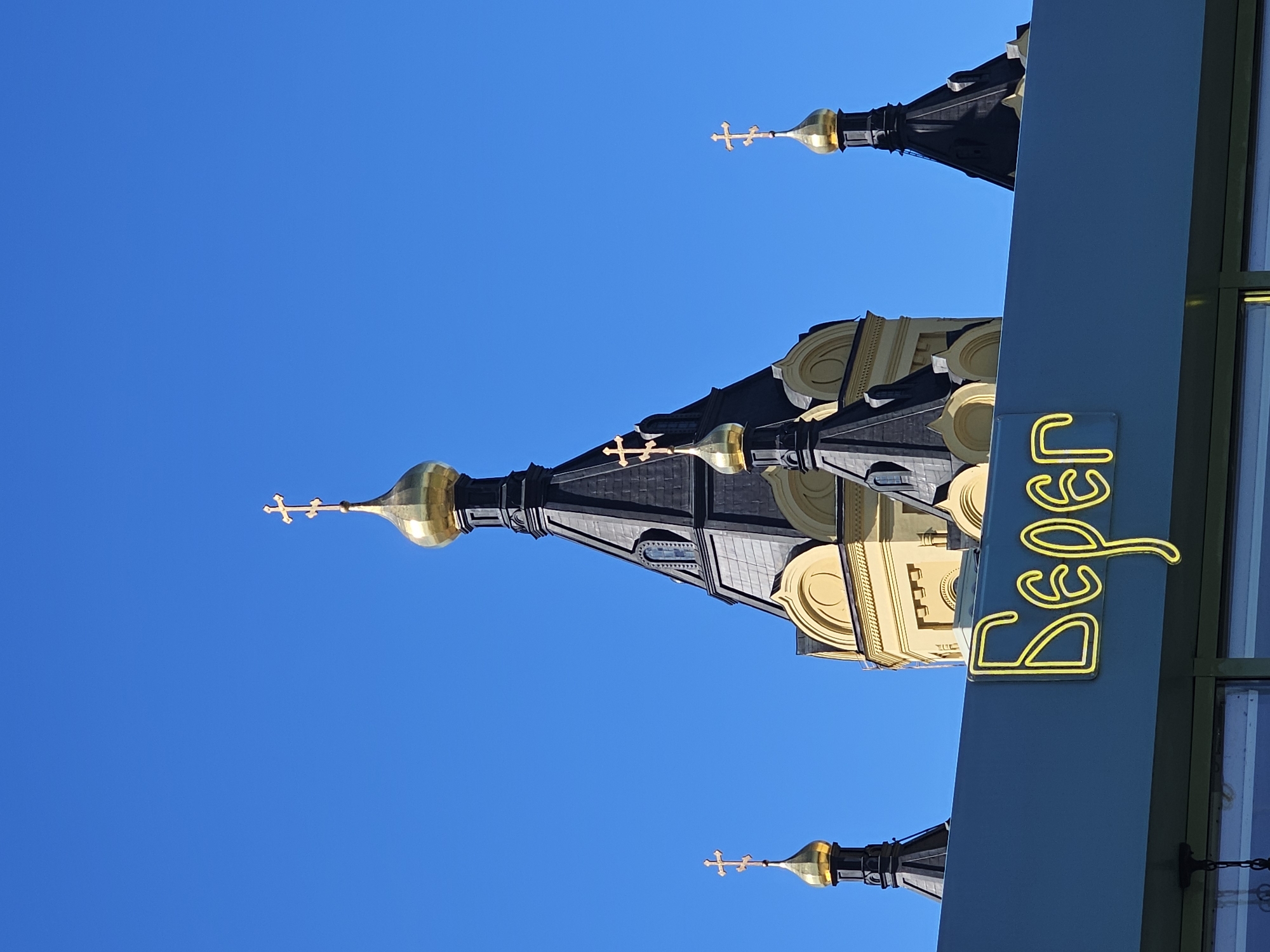 The image shows a view of the Nizhny Novgorod Kremlin's Alexander Nevsky Cathedral, a prominent landmark in Nizhny Novgorod, Russia. The bright blue sky serves as a backdrop to the cathedral's golden domes and black spires. In the foreground, a blue sign with neon yellow Cyrillic lettering, which reads "Берег" (Bereg), is visible. This suggests the presence of a building or establishment with this name, potentially a cafe, shop, or restaurant, located near the Kremlin. The architecture of the cathedral is a characteristic example of Russian Revival style. There are no people visible in the image. The lighting indicates it is daytime, likely with clear weather.