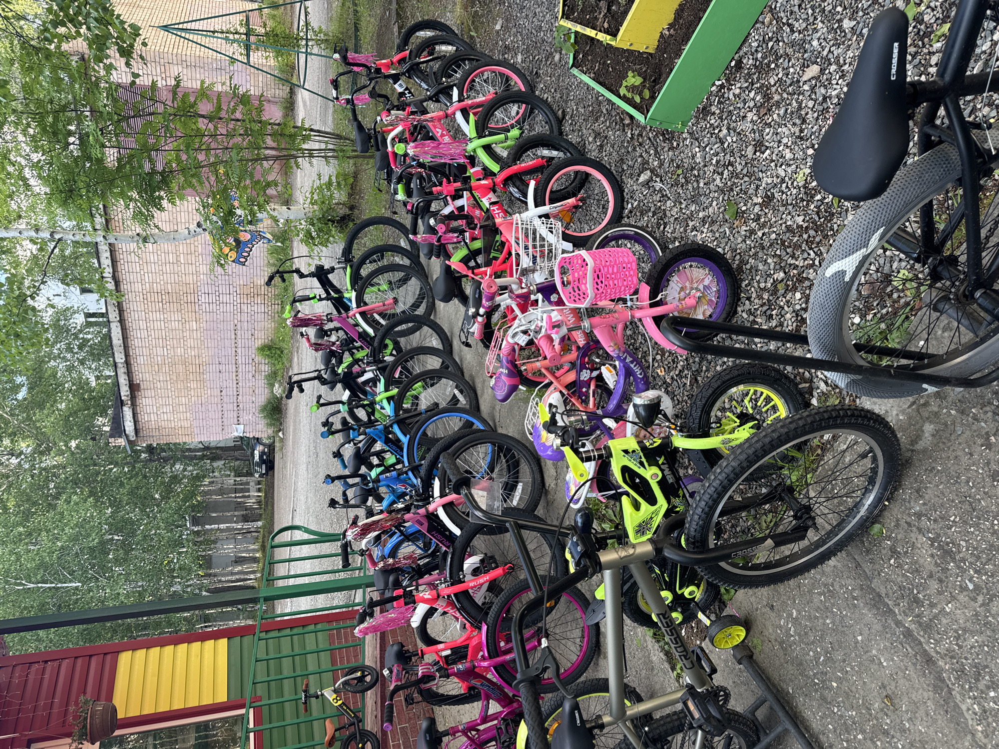 In the city of Tynda, Russia, a collection of bicycles is neatly parked on a gravel surface in front of a building with red and yellow siding, bordered by a green railing. The bicycles, varying in size and color, include several pink and lime green models, some with training wheels and decorative streamers on the handlebars. A small balance bike with a brown seat is positioned on the left side of the frame. In the background, a light brick building stands, with trees and foliage surrounding the area. A portion of a green metal structure, possibly playground equipment, is visible on the right. The scene appears to be an outdoor storage area or a rental station for bicycles.