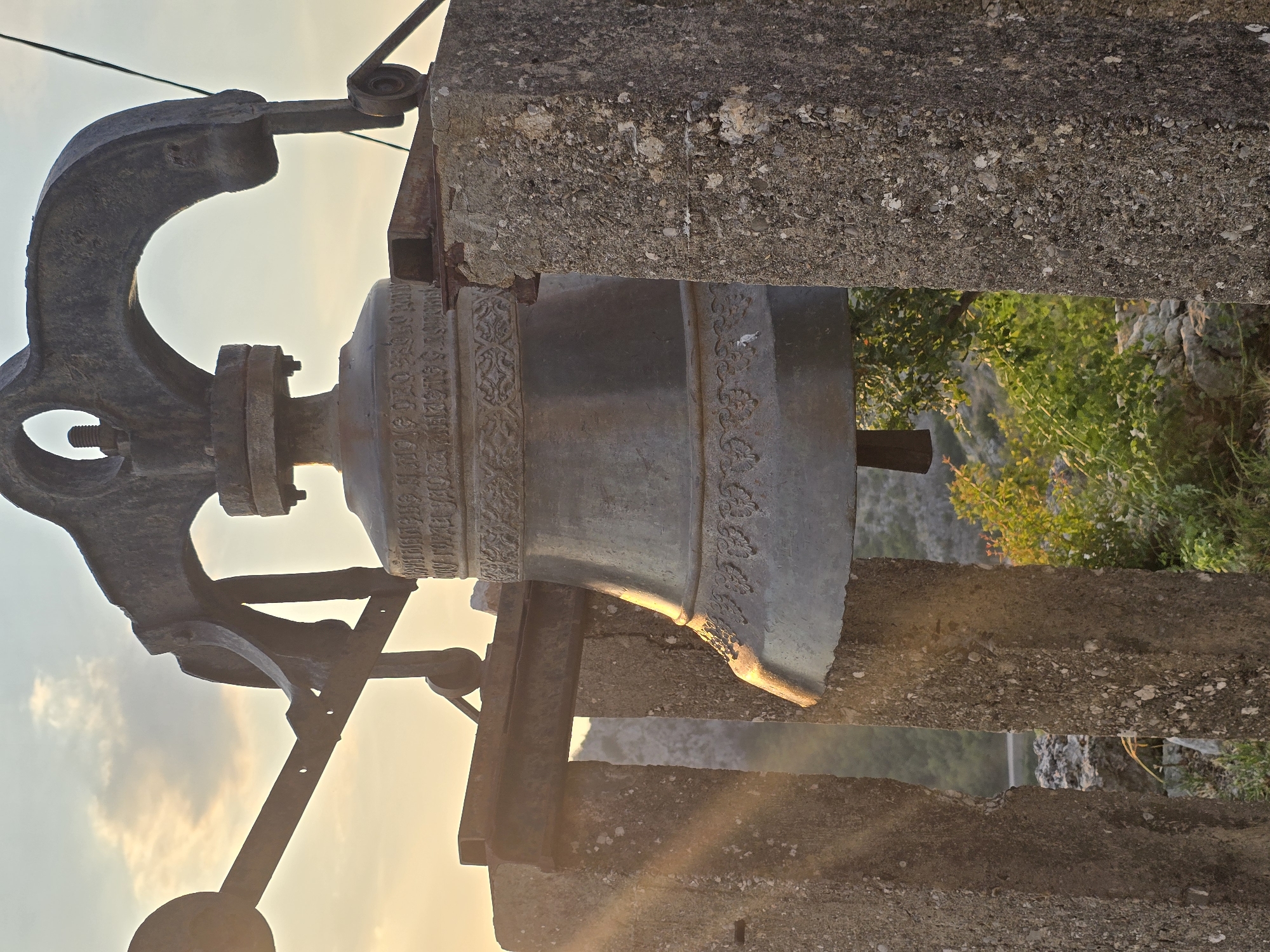 The image shows a large, weathered bronze bell mounted on a rustic metal frame supported by two rough-hewn concrete pillars. The bell itself is ornate, with decorative bands around its upper and lower sections, and appears to have some form of inscription on its upper rim. Behind the bell, sunlight streams through the clouds, creating a warm, golden glow and lens flare effect. The background reveals a rocky, mountainous landscape with sparse vegetation, suggesting an outdoor location. The scene conveys a sense of age and permanence. The city of Gornja Vrbica, Montenegro, is not visible in this image.