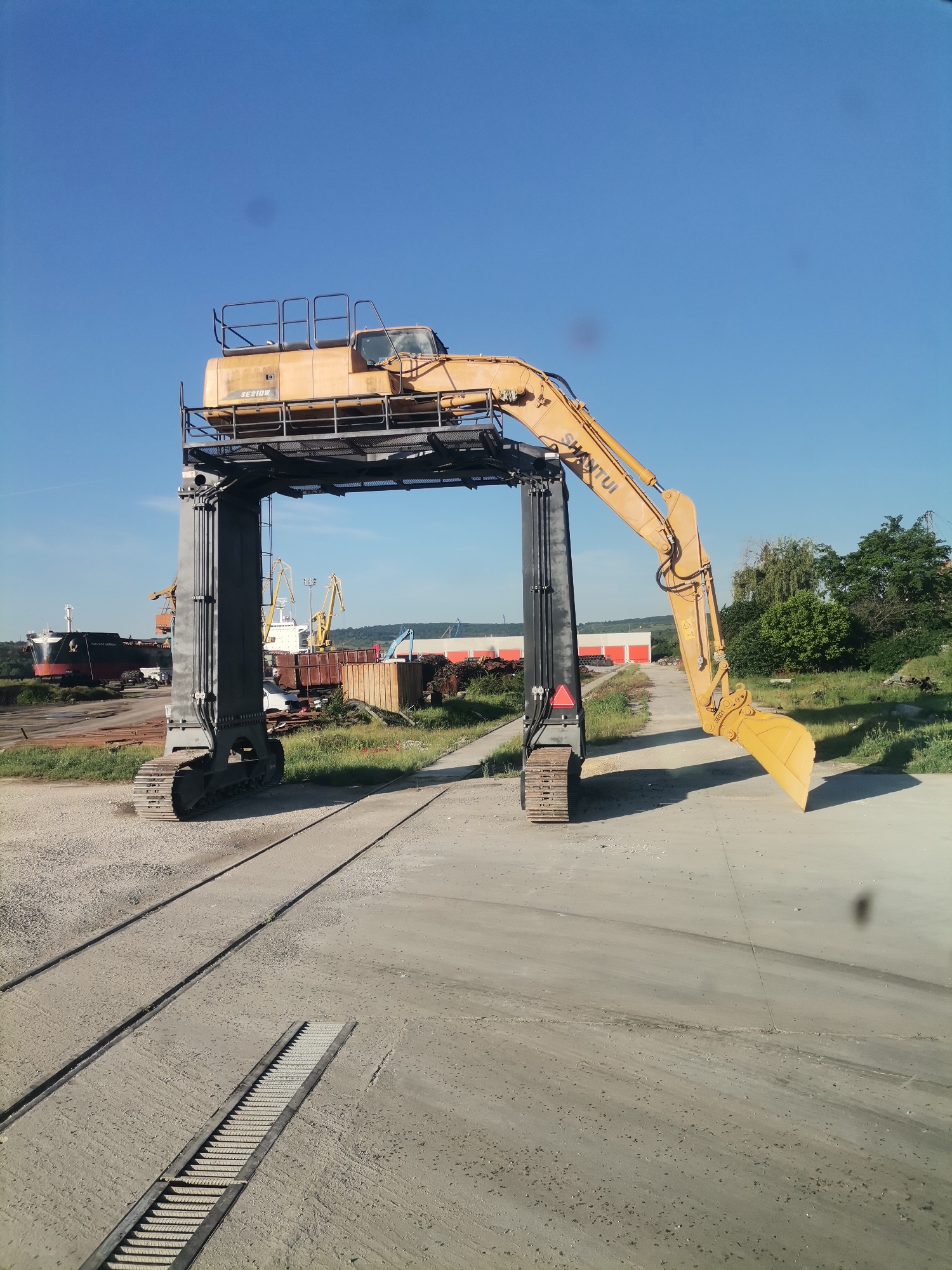 The image shows a large, yellow Shantui SE210W excavator converted into a gantry crane. The excavator is positioned atop two vertical columns supported by tank treads, creating an archway. The excavator's arm and bucket are extended downward on the right side. The entire structure is placed on a concrete and gravel surface with visible train tracks. In the background, there are industrial scenes, including cargo ships, cranes, storage containers, and piles of scrap metal. Patches of vegetation are visible near some red storage containers. The scene is located near Ezerovo, Bulgaria, based on geolocation clues.
