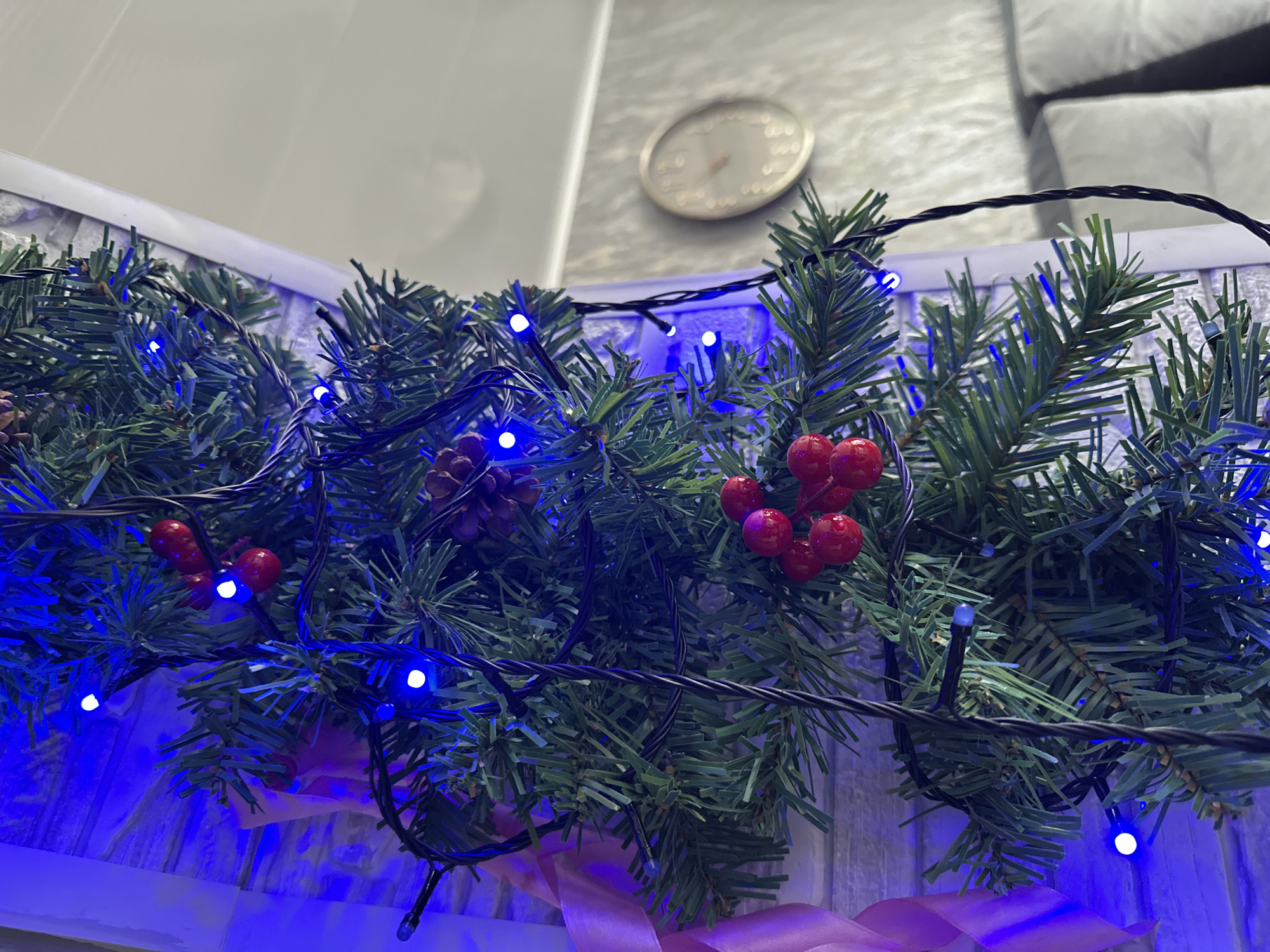 An artificial evergreen garland, adorned with blue LED string lights, clusters of red artificial berries, and small natural pinecones, is displayed vertically on a light-colored, textured surface, possibly wood paneling or white brick-patterned wallpaper. The blue LED lights are irregularly spaced along a black wire, casting a distinct blue glow on the immediate background. A pale pink ribbon is partially visible on the far left, woven into the garland. A thin white trim or frame delineates the right edge of the garland's mounting surface. In the blurred background to the right, a circular analog wall clock with a silver-toned bezel and a light-colored face is mounted on a light gray wall. Below and to the right of the clock, a section of light gray upholstered furniture is visible. The scene is indoors, within a residential setting in the City of Unknown, Unknown. No individuals are present.