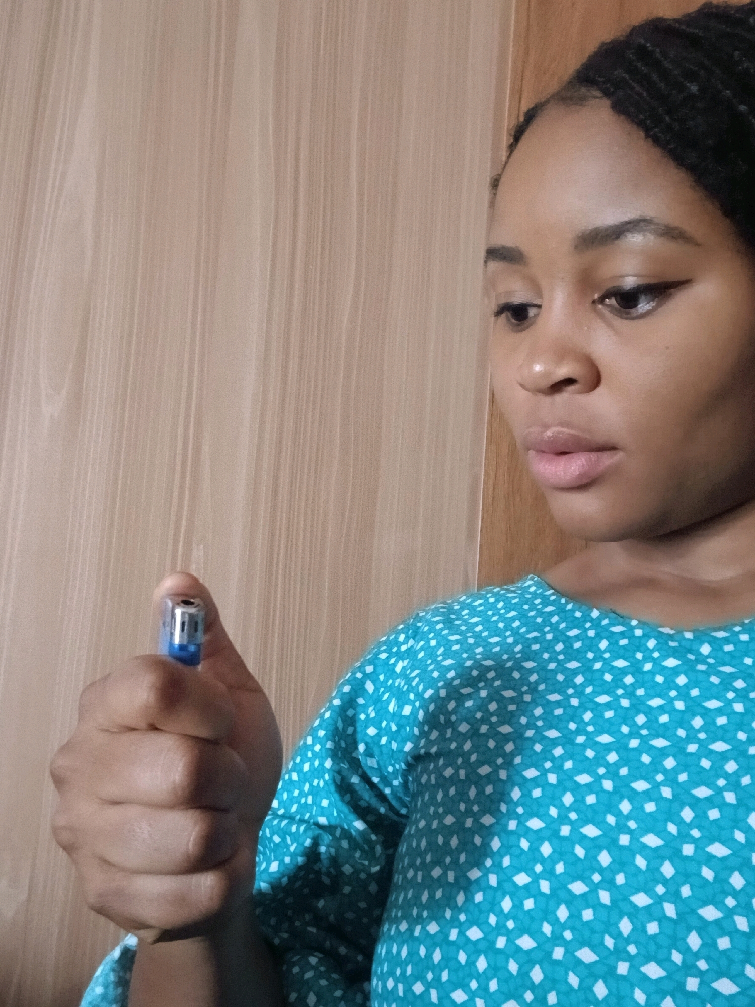 A medium shot of a young Black woman with braided hair, wearing a teal shirt with a white diamond pattern, is shown from the chest up. She is holding a blue and silver lighter in her right hand, with her thumb and index finger around it. The background is a light brown, vertically striped wooden surface. The woman is looking downwards and to her left, with her mouth slightly open. The scene appears to be indoors.